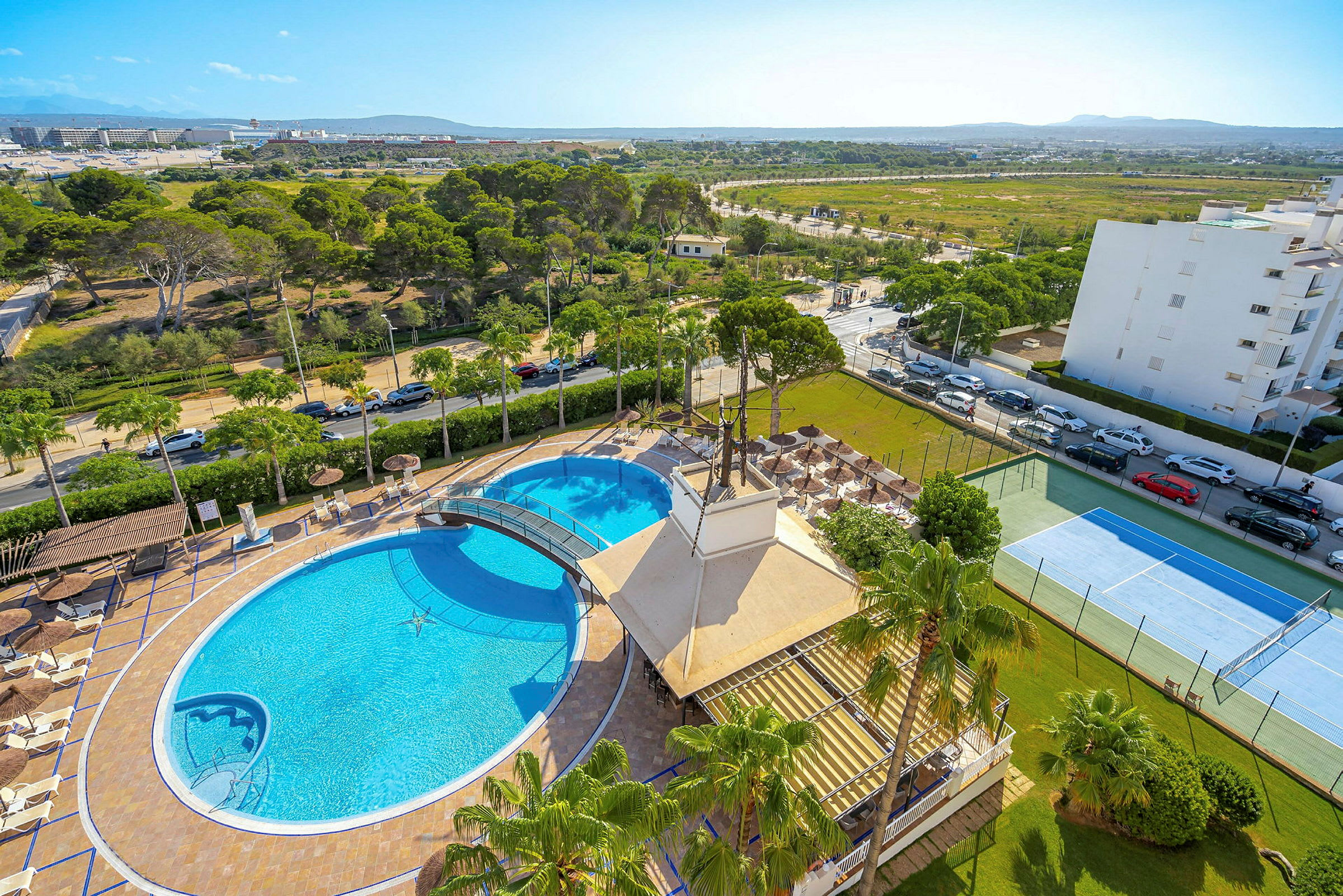 Utsikt över hotellområde med pool, parasoller, grönytor och tennisbana. (AI-genererad text)