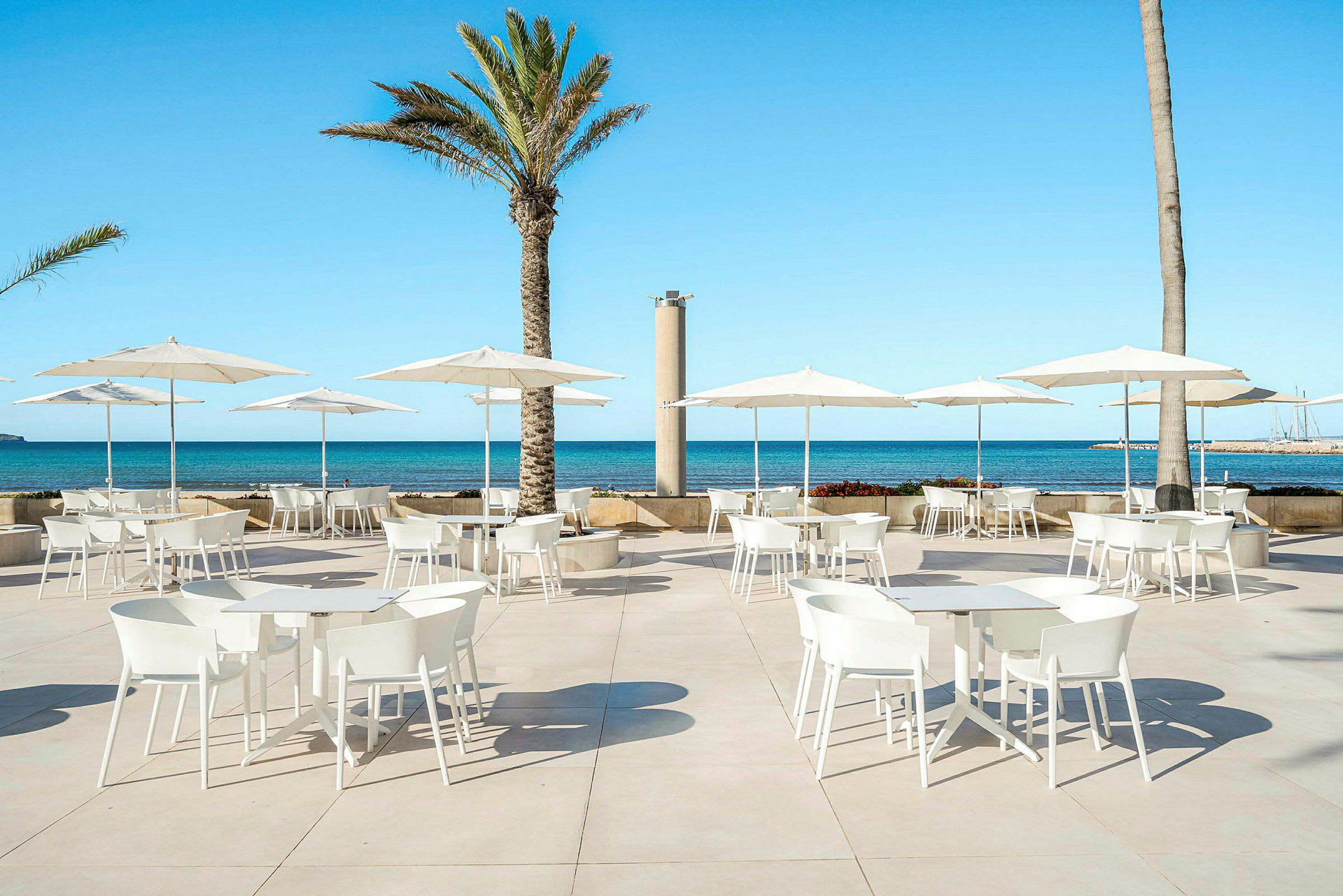 Vitmålade bord och stolar med parasoller på en terrass vid havet. (AI-genererad text)