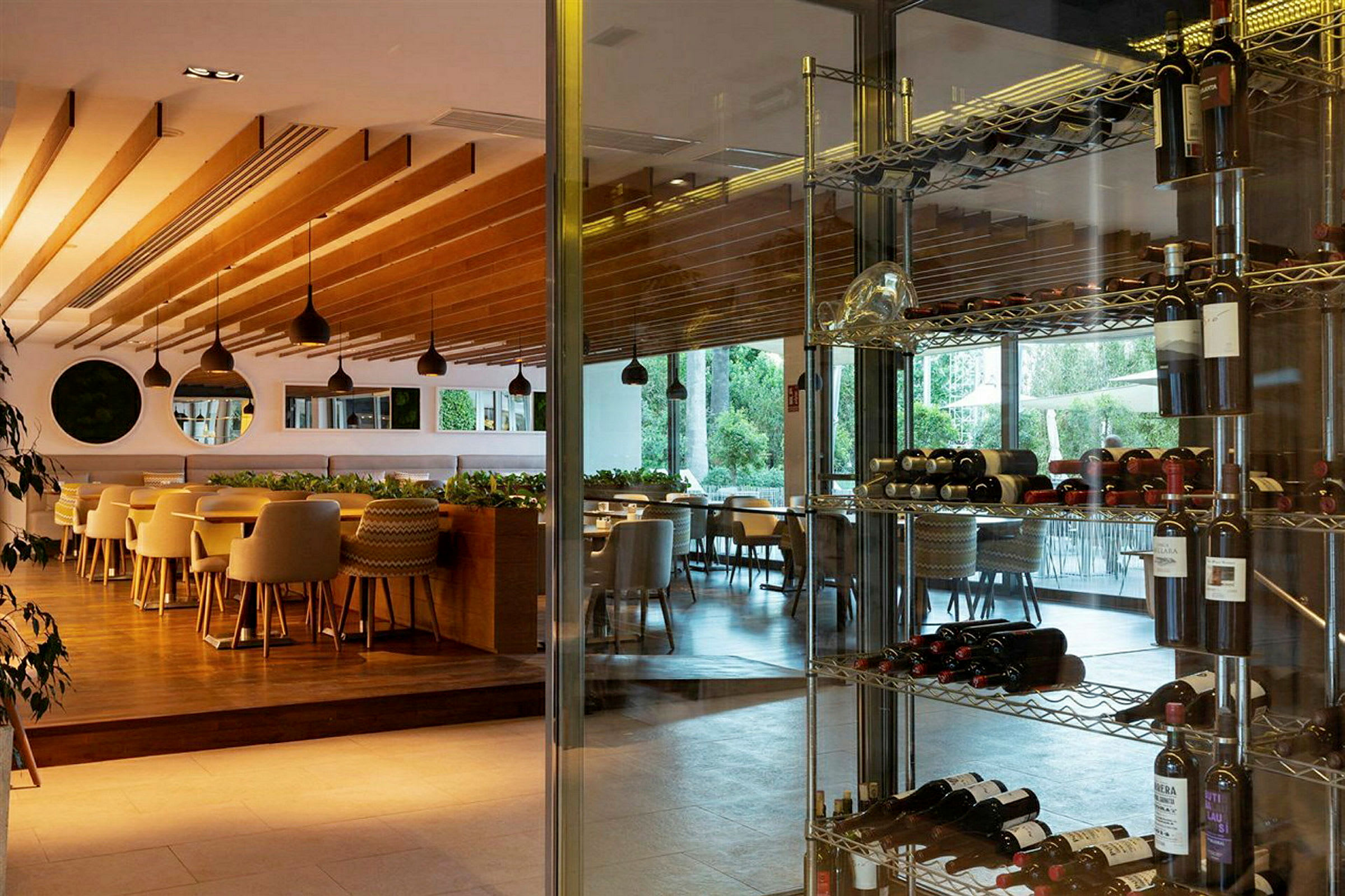 En ljus restaurang med trädetaljer på taket, stora fönster med utsikt mot grönska, och vinflaskor på metallhyllor. (AI-genererad text)