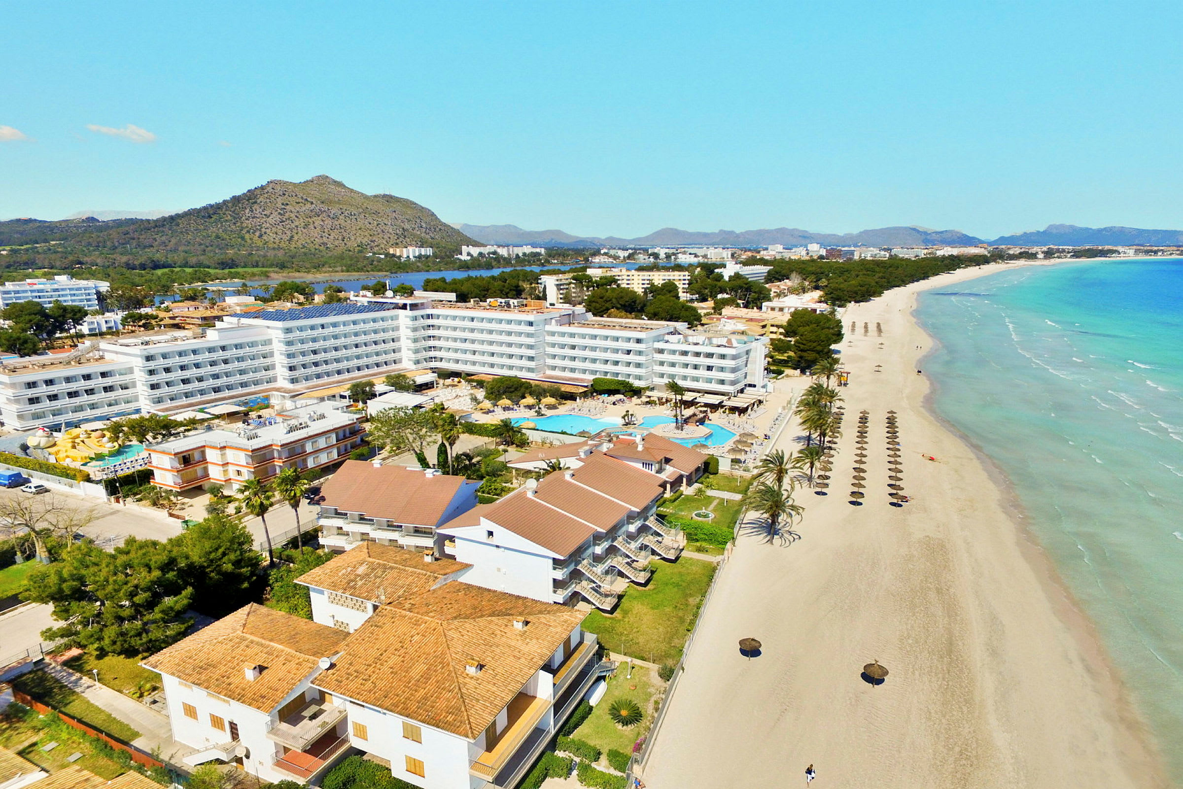 Hotellbyggnader och poolområde vid en lång sandstrand med parasoller samt berg och grönska i bakgrunden. (AI-genererad text)