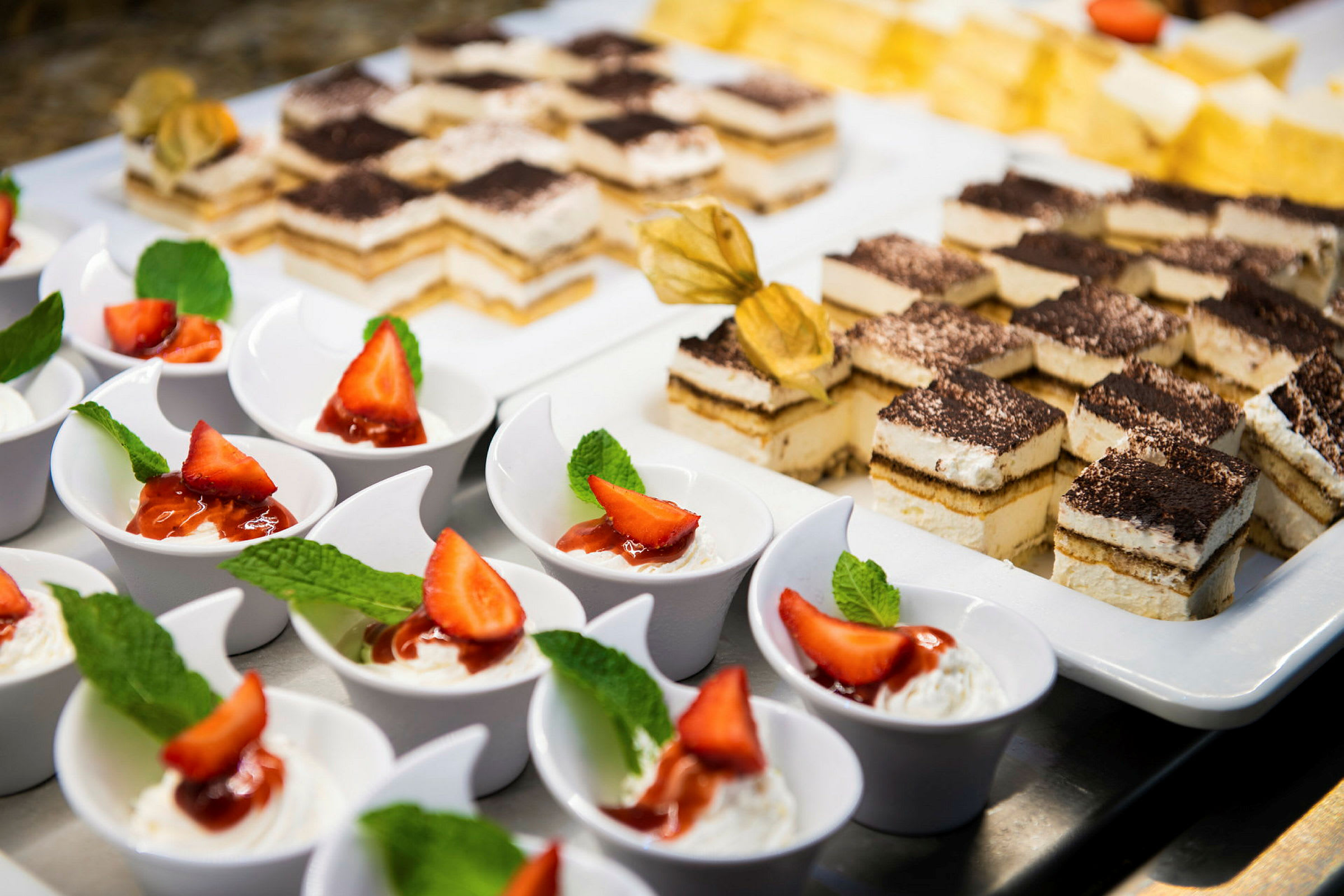 En buffé med individuella desserter, bland annat vit skål med grädde, jordgubbar och mynta samt fyrkantiga bakverk med frosting. (AI-genererad text)