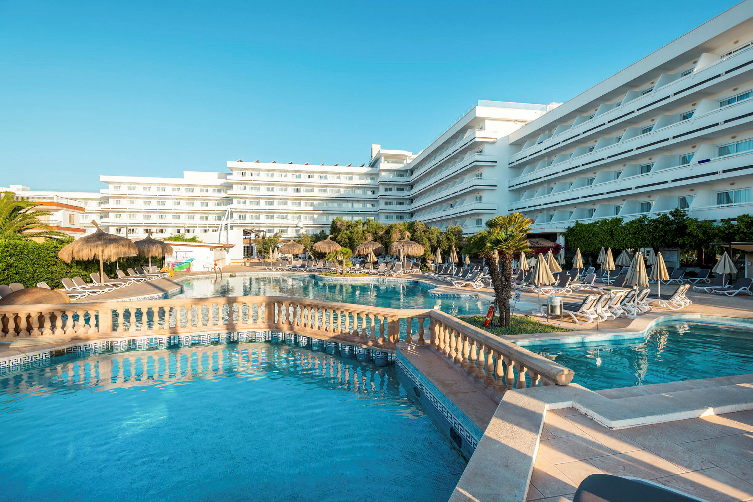 Stor hotellpool omgiven av solstolar, parasoller och gröna växter mot en bakgrund av vita byggnader. (AI-genererad text)