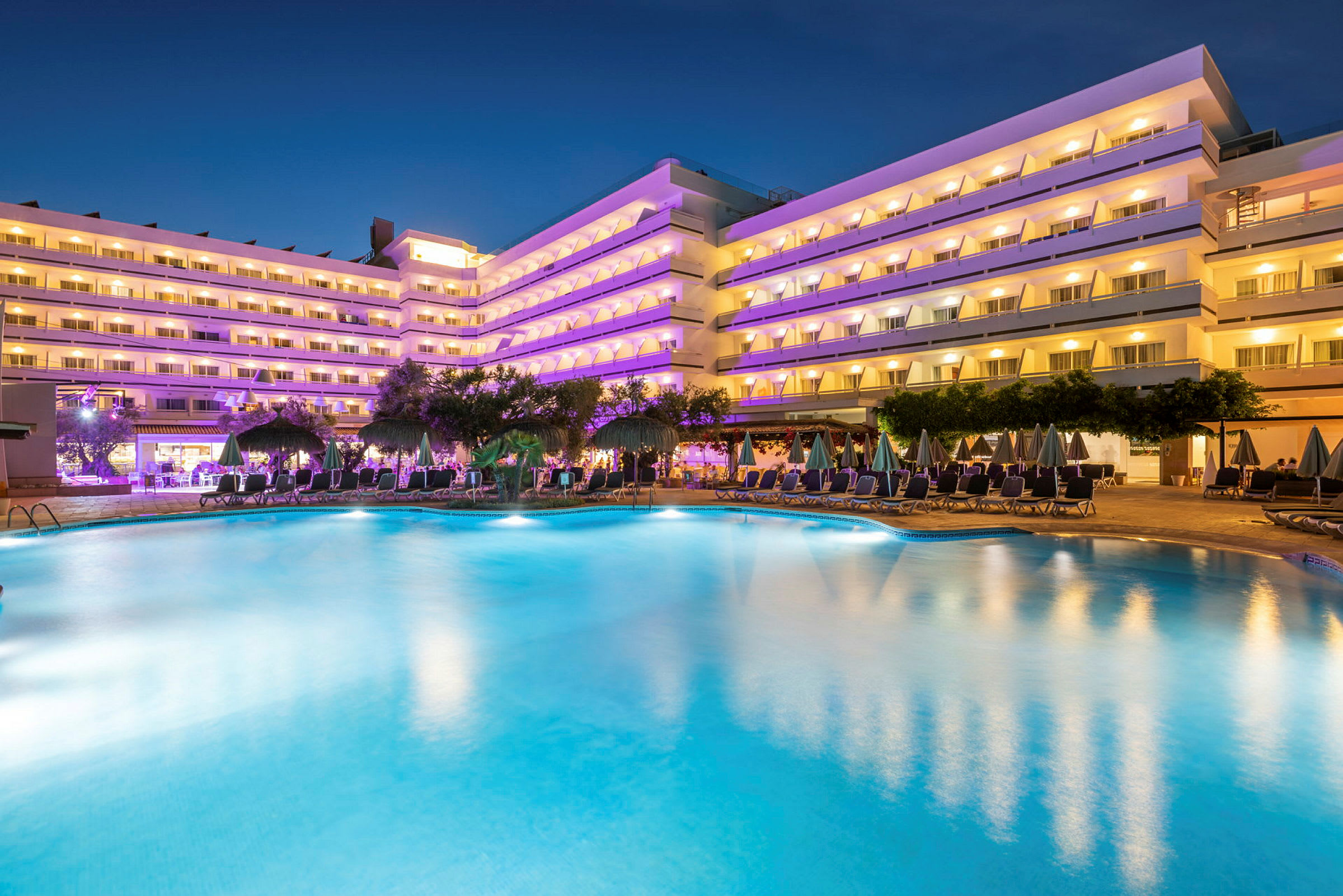 Moderna hotellbyggnader med upplysta balkonger synliga vid en stor pool omgiven av solstolar och parasoller under kvällen. (AI-genererad text)