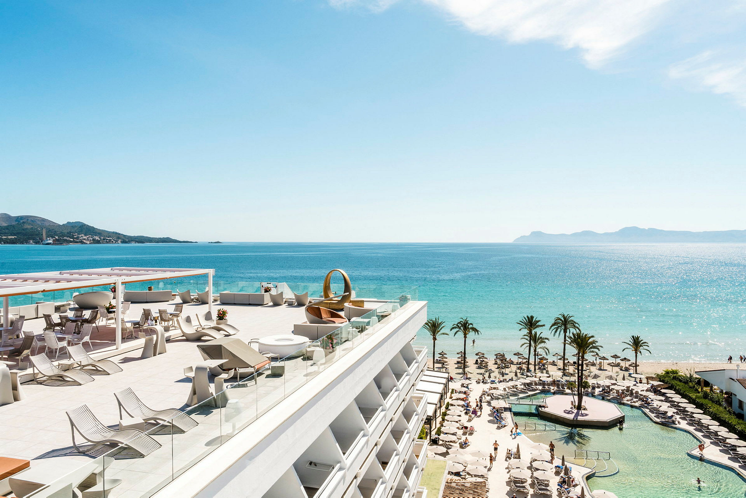 Visar en högt belägen terrass med solstolar och bord, kustnära utsikt med strand, palmer och hav. (AI-genererad text)