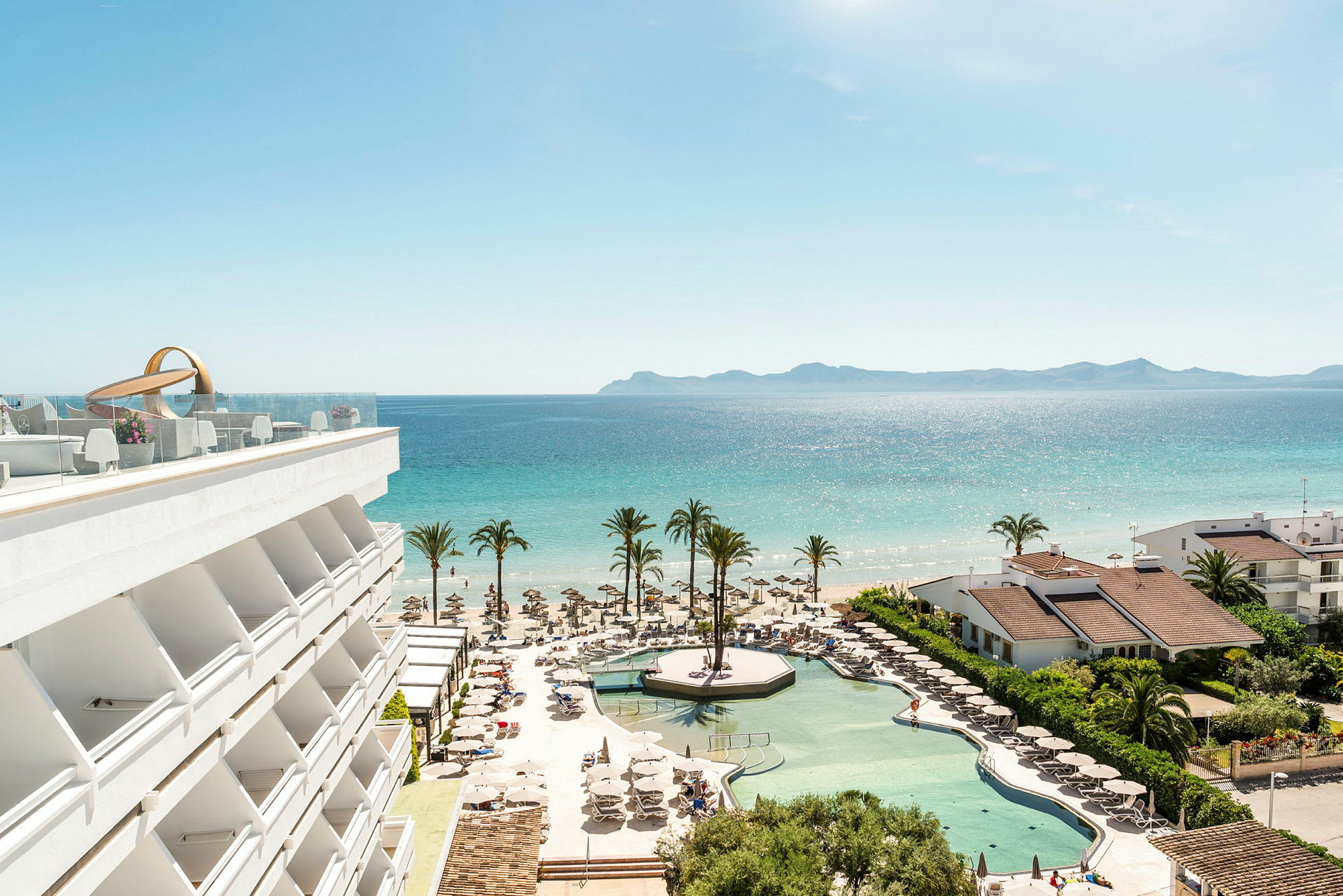 En utsikt över hotellpool, strand och hav med palmer och byggnader. (AI-genererad text)