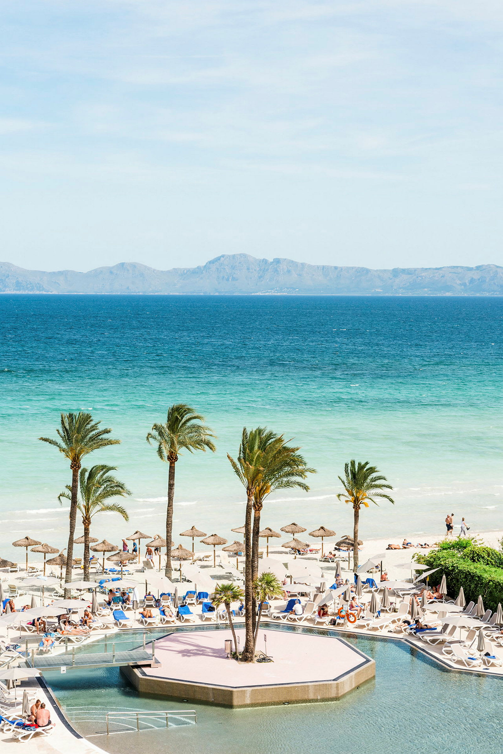 Utsikt över en pool och stranden med solstolar, parasoller och palmer vid vattnet. (AI-genererad text)