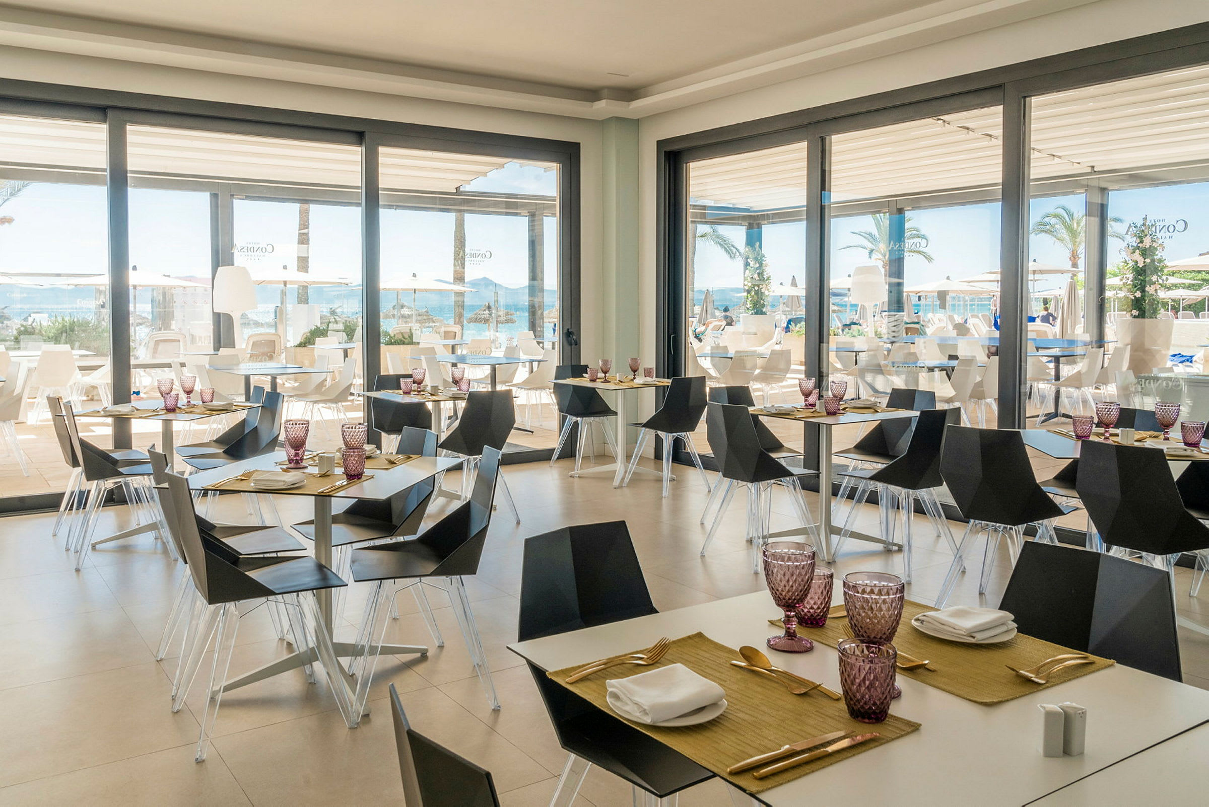 Restaurang med moderna möbler och dukade bord vid stora glasfönster med utsikt mot pool och hav. (AI-genererad text)