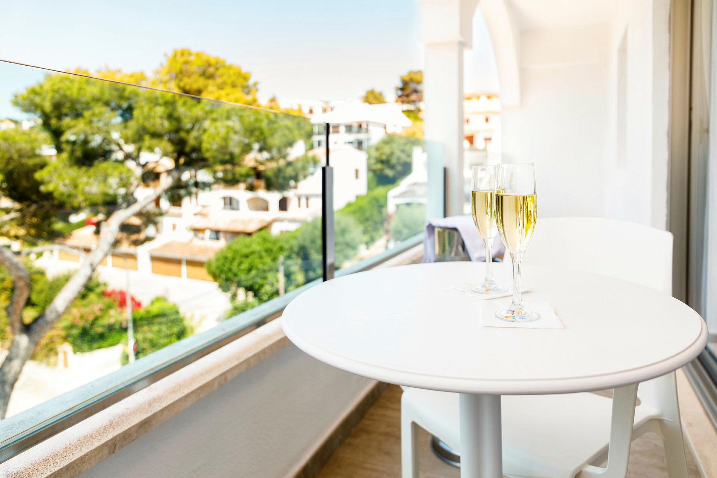 Ett vit bord med två glas champagne vid en balkong med utsikt över en grönskande trädgård och hus i bakgrunden. (AI-genererad text)