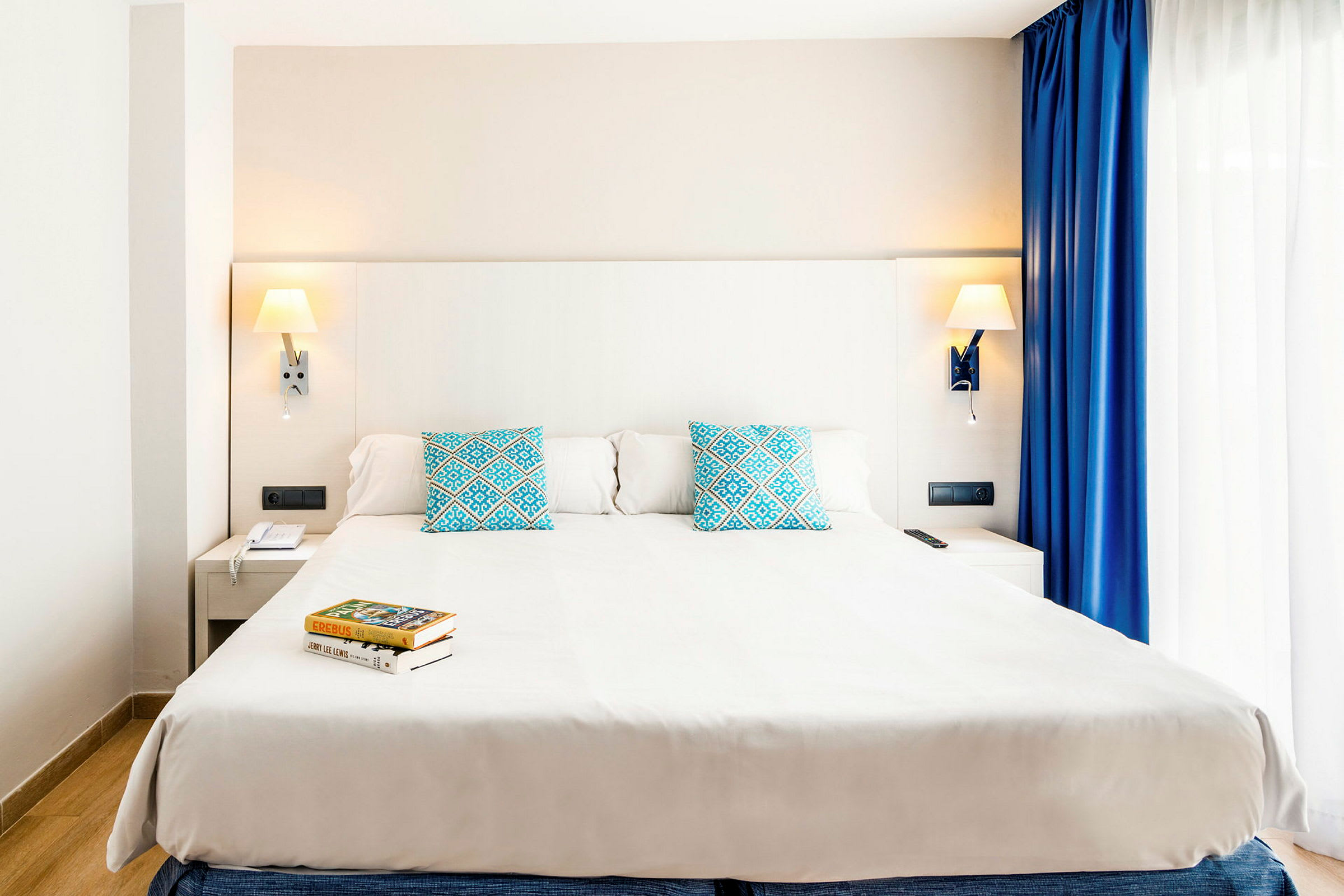 Ett modernt hotellrum med en stor dubbelsäng, två blå prydnadskuddar med mönster, och läslampor på väggen. Bredvid sängen finns ett nattduksbord och ett fönster med vita och blå gardiner. (AI-genererad text)
