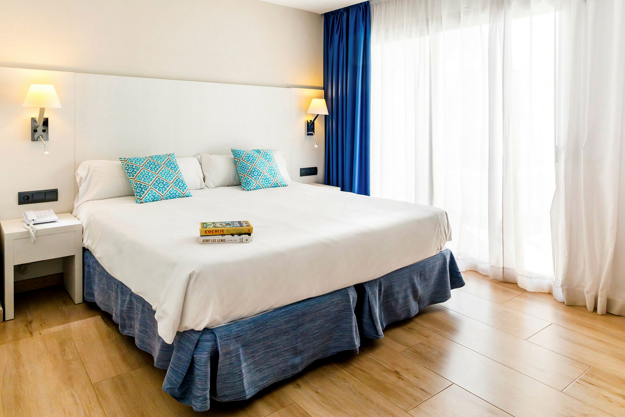 Ett hotellrum med en bred säng, vit sänggavel, blå kuddar och gardiner samt en ljus träparkett. (AI-genererad text)