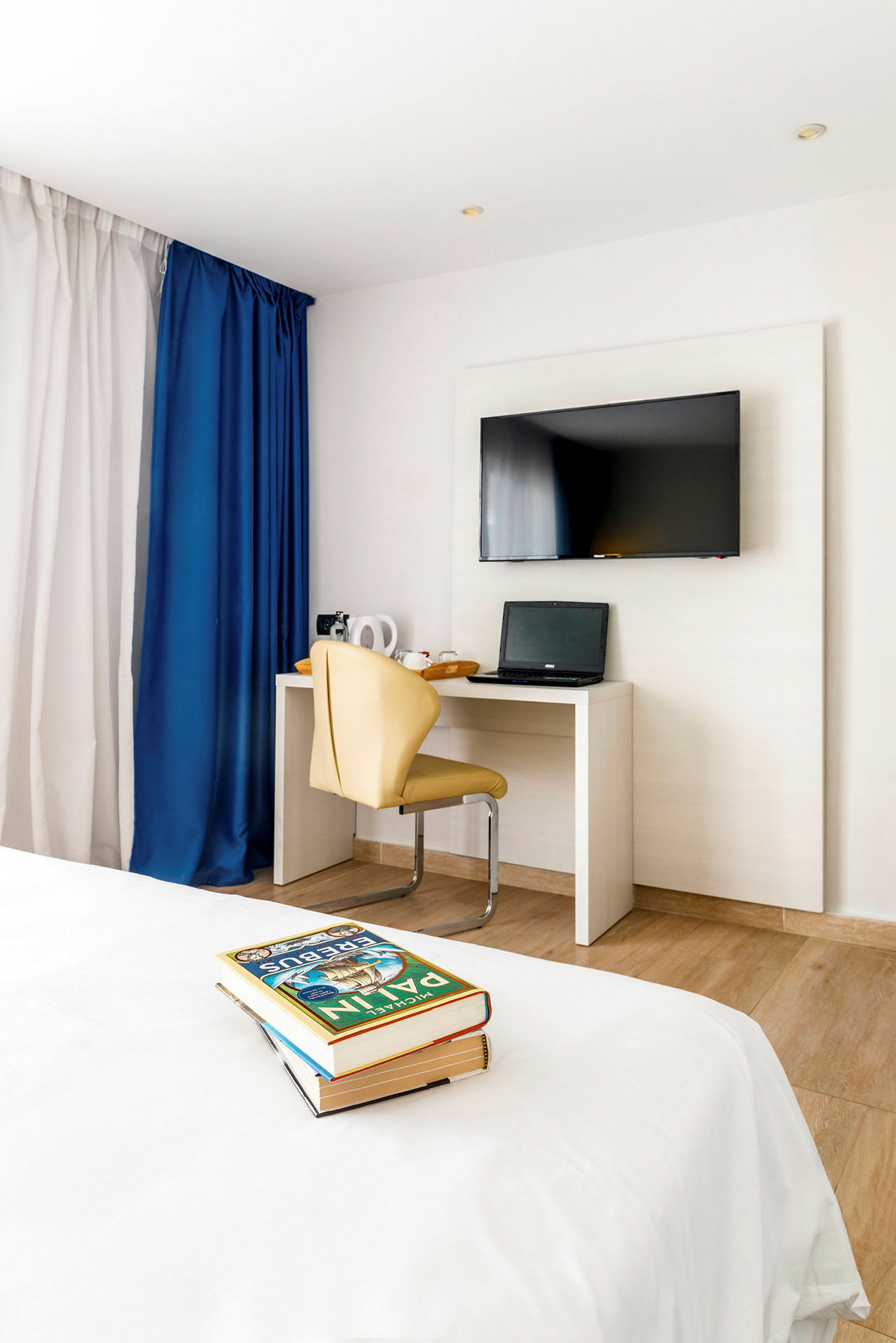 Ett hotellrum med en platt-TV på väggen, ett skrivbord med en laptop och en beige stol. I förgrunden syns en säng och en hög med böcker. (AI-genererad text)