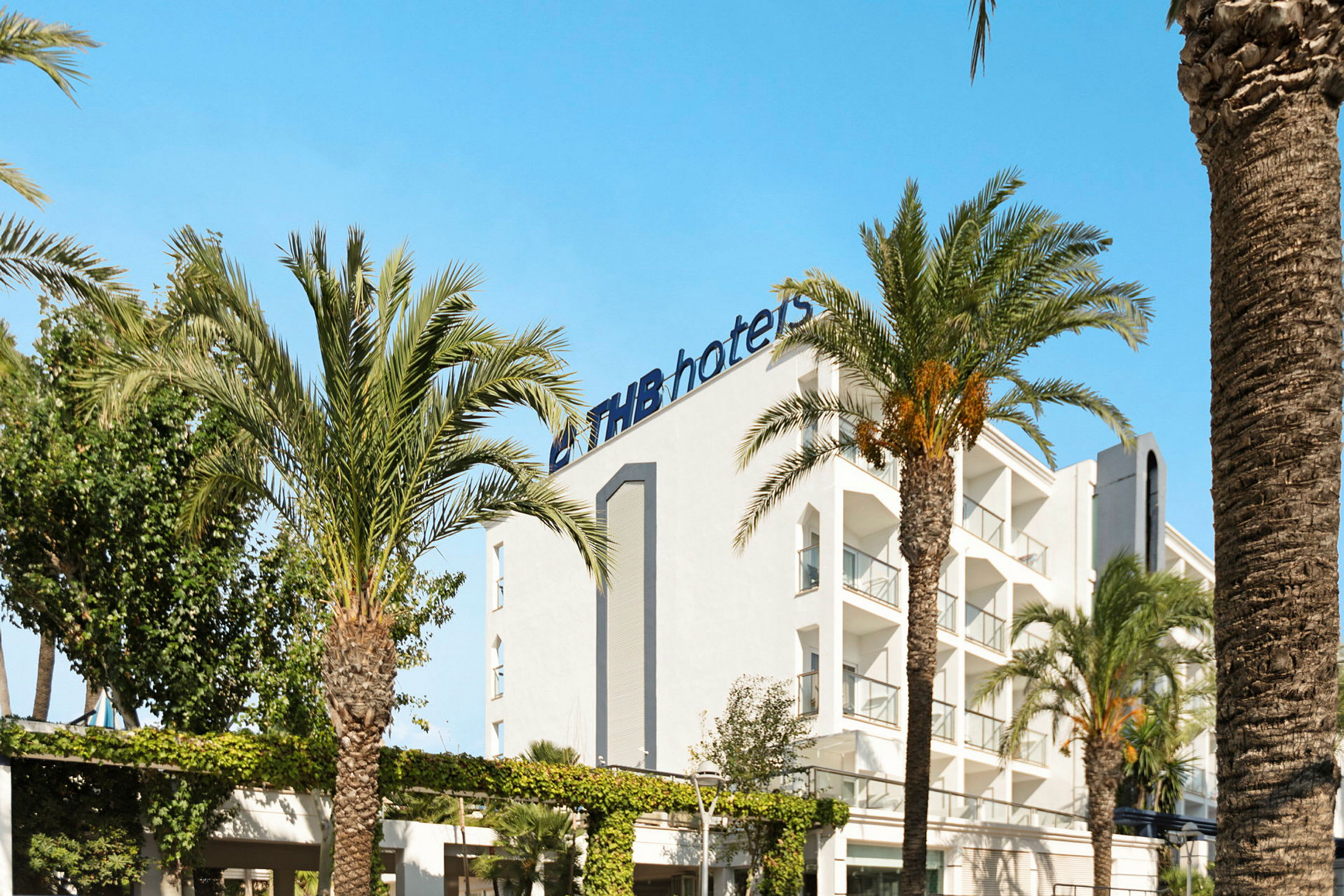 Vit byggnad med logotypen 'THB hotels' omgiven av palmer under en klarblå himmel. (AI-genererad text)