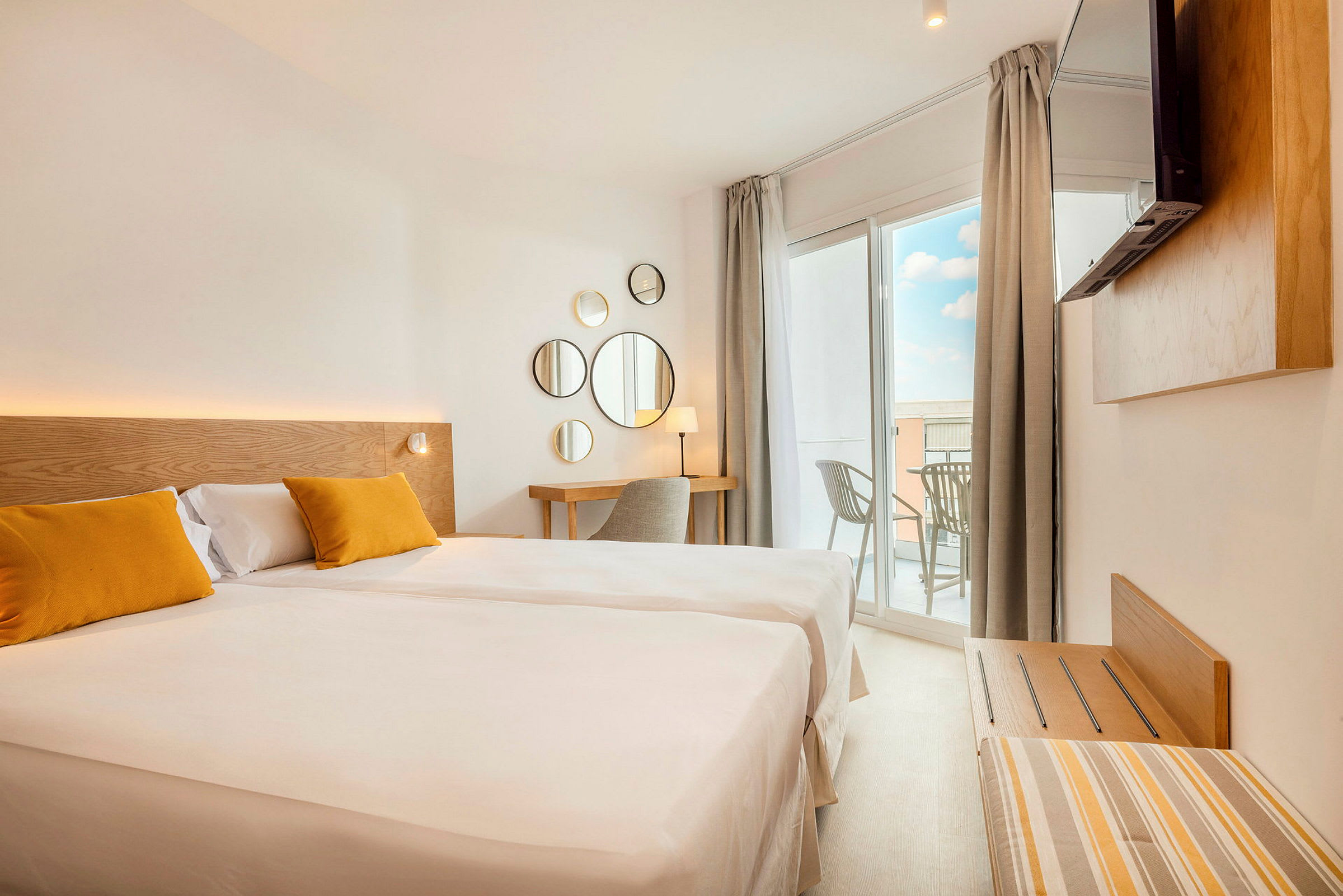 Ett ljust hotellrum med en stor säng, två gula kuddar, en skrivbordshörna och en balkong med utsikt. (AI-genererad text)
