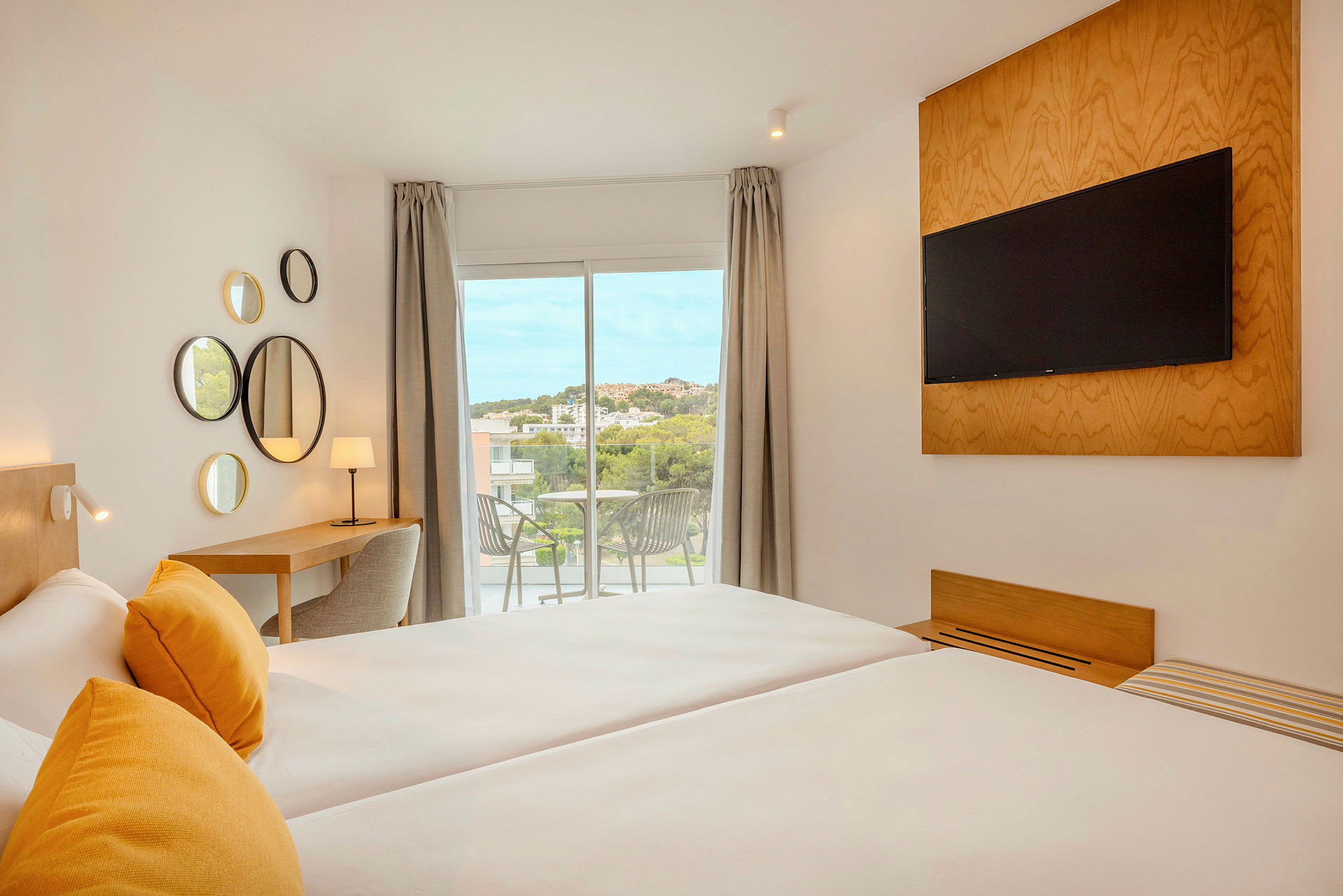 Ett modernt hotellrum med två sängar, gula kuddar, en TV monterad på väggen, ett skrivbord med lampa och en balkong med utsikt. (AI-genererad text)