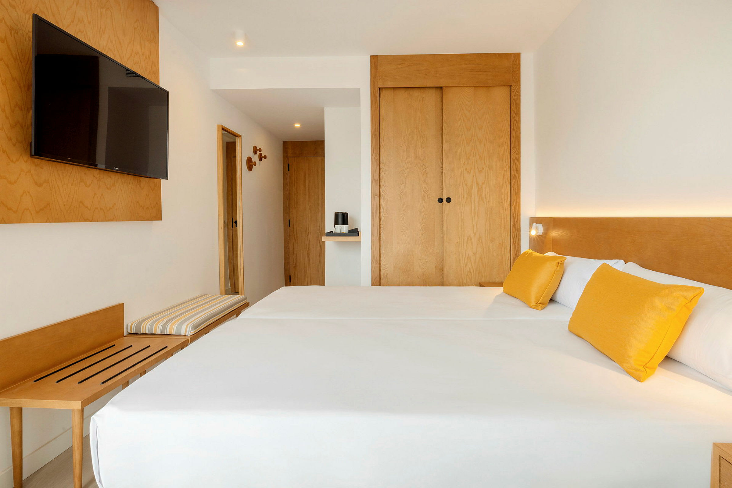 Ett hotellrum med modern inredning, två enkelsängar med vita sängkläder och gula kuddar, en väggmonterad TV och trämöbler. (AI-genererad text)