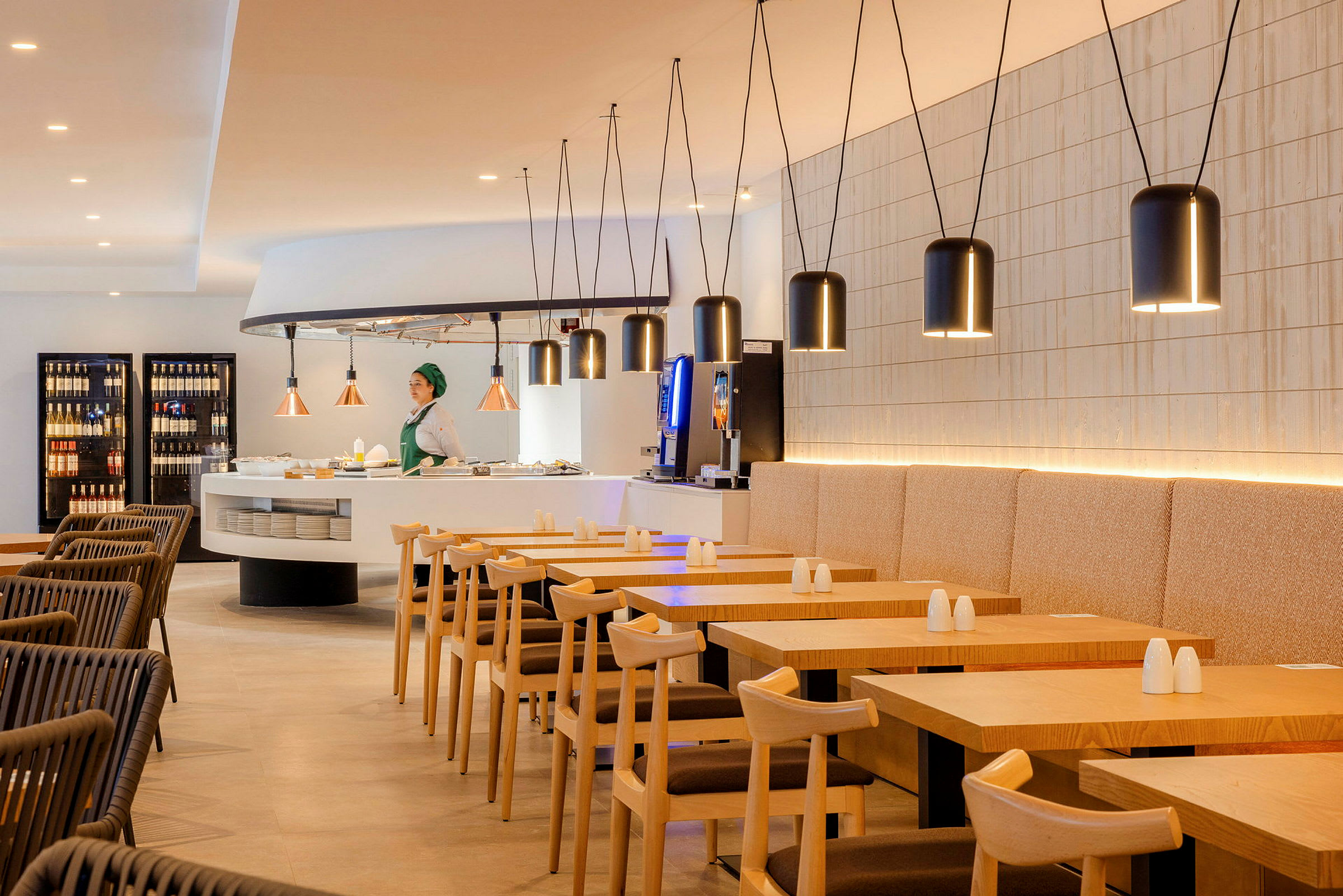 Bild av en modern restaurangmiljö med träbord och stolar. En person står vid en serveringsdisk omgiven av hängande lampor. (AI-genererad text)