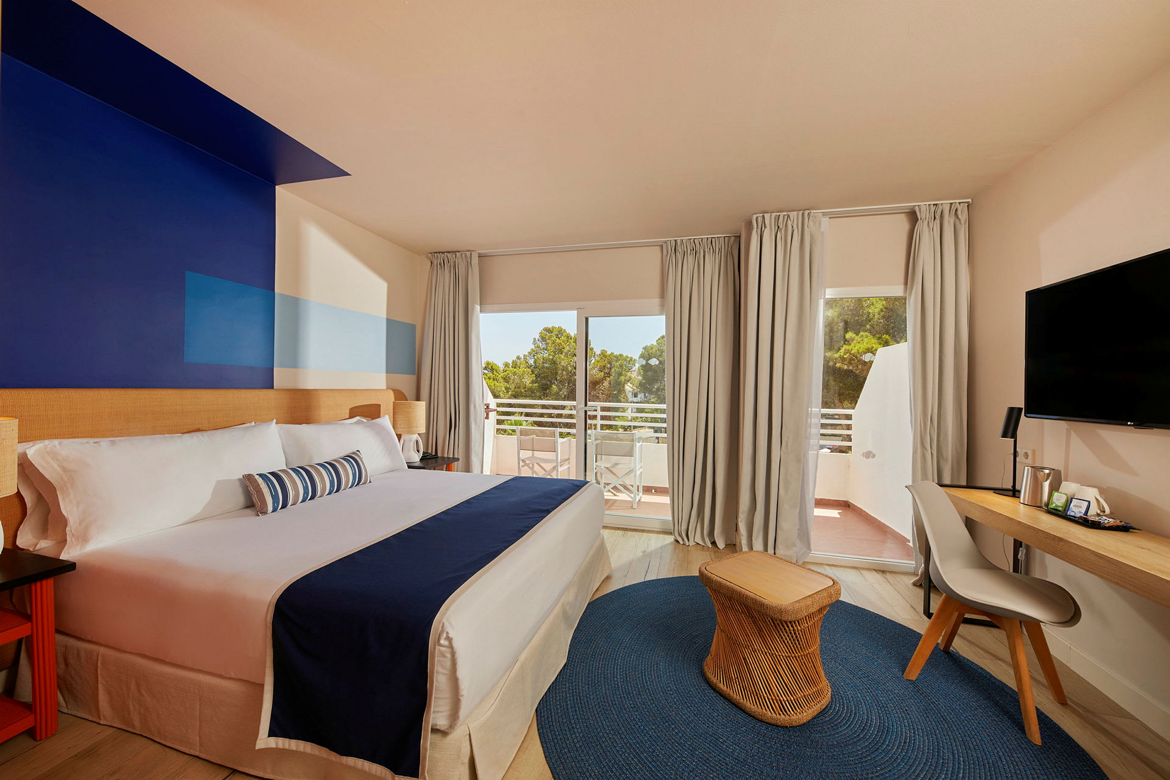 Hotellrum med dubbelsäng, skrivbord och stol, samt balkong med utsikt över träd. (AI-genererad text)