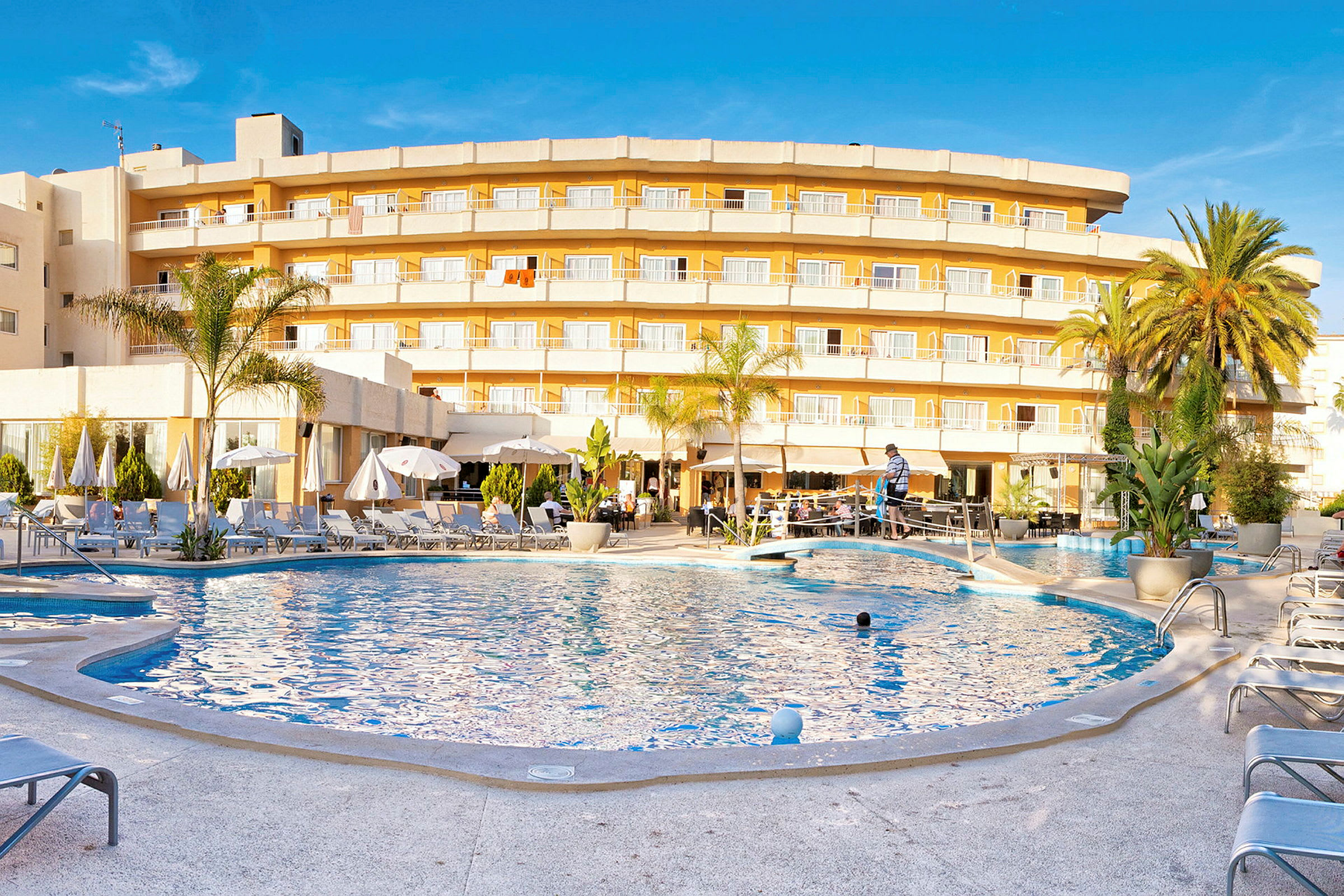 Hotell med gul fasad och balkonger framför en swimmingpool omgiven av solstolar. (AI-genererad text)