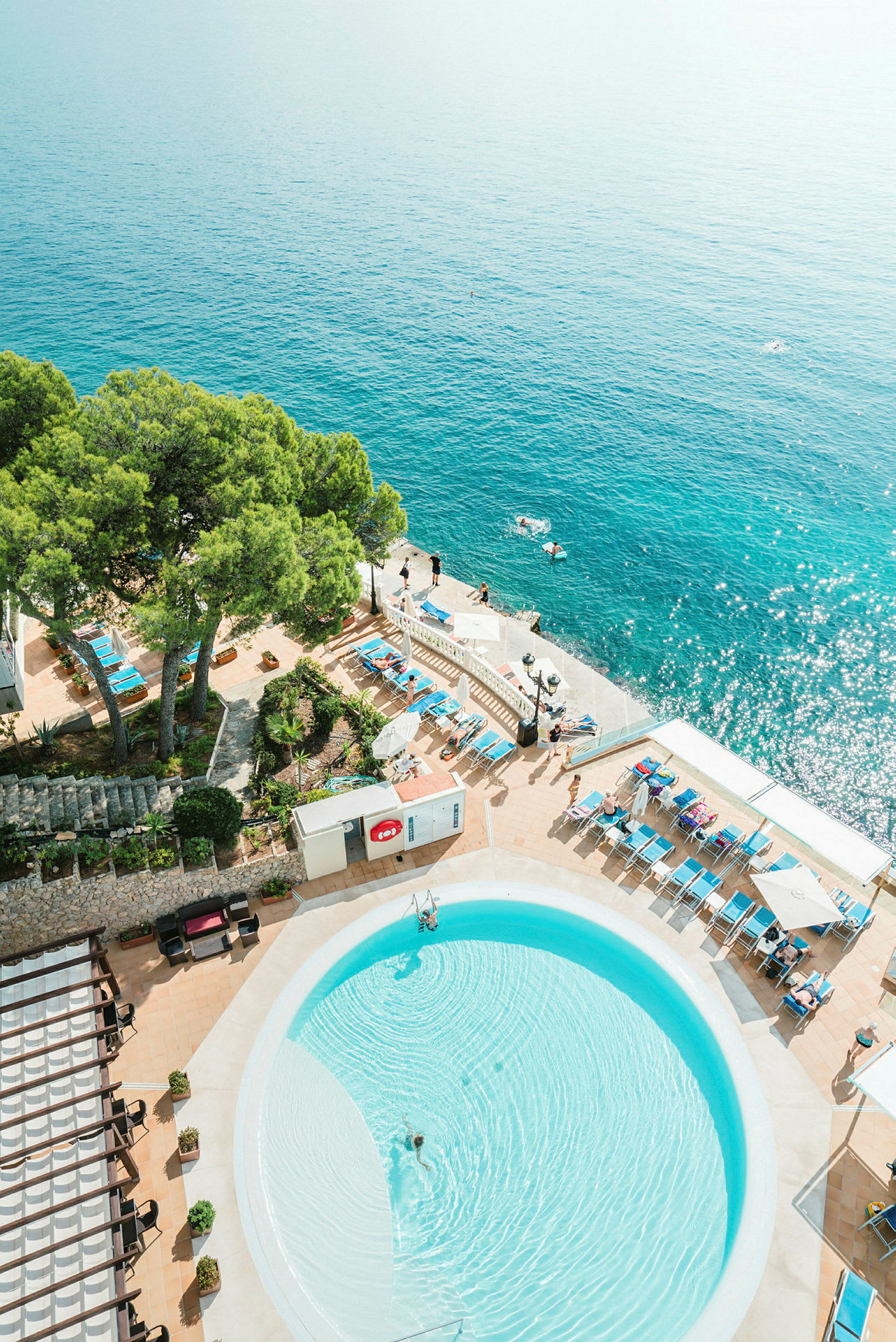 Vy ovanifrån på en pool omgiven av solstolar och gröna träd med havet i bakgrunden. (AI-genererad text)