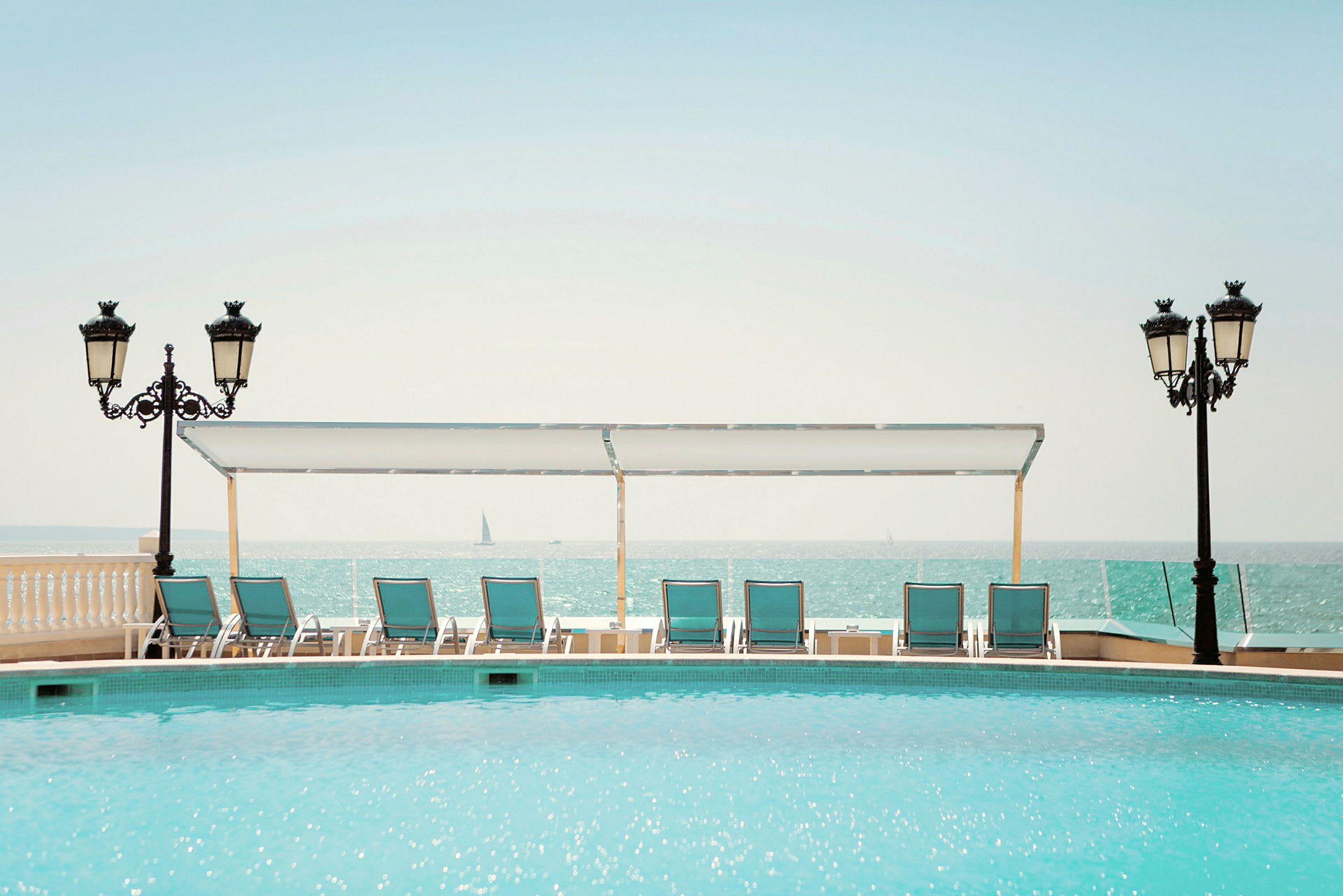 En pool med solstolar och parasoll i förgrunden, samt havet och segelbåtar i bakgrunden. (AI-genererad text)