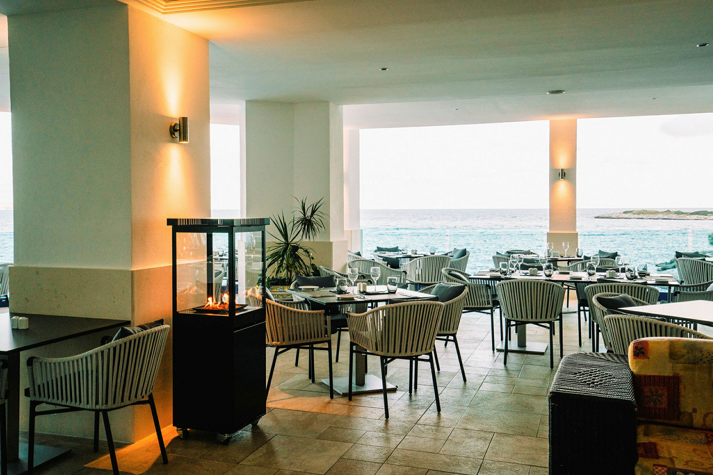 En restaurang med utomhusutsikt över havet, möblerade med bord och stolar och dekorerade med en liten eldstad. (AI-genererad text)