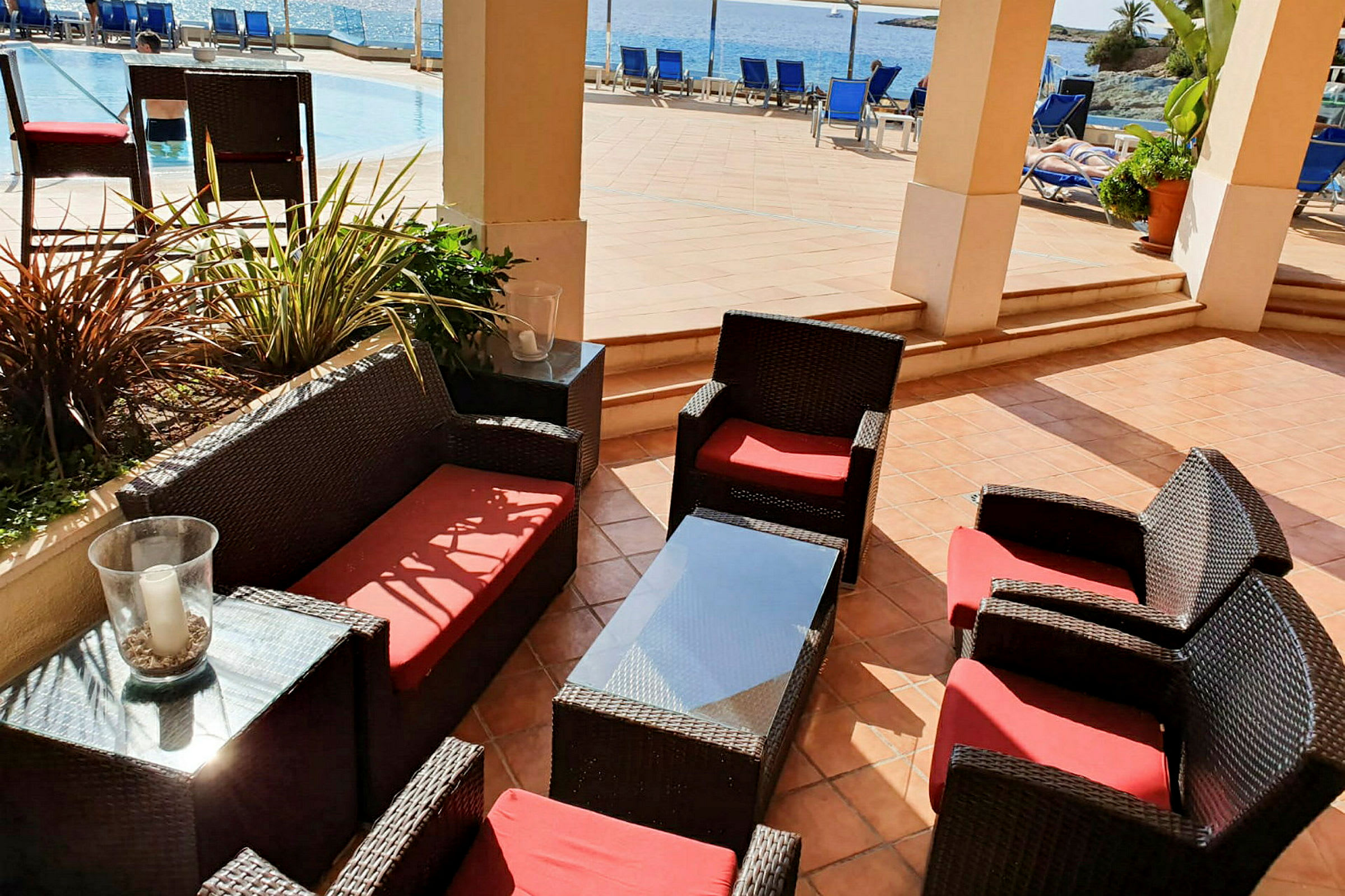 Utemöbler med röda dynor på en terrass nära pool och hav. (AI-genererad text)