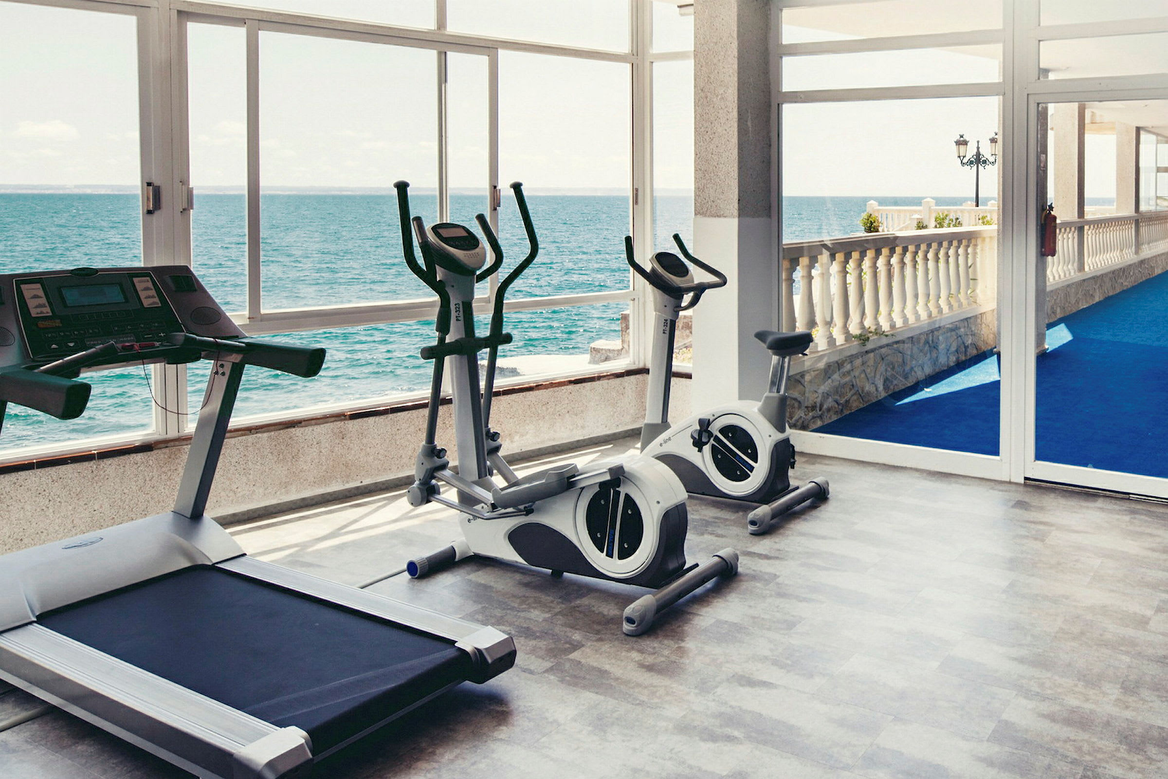 Gym med träningsutrustning och havsutsikt genom glasvägg. (AI-genererad text)