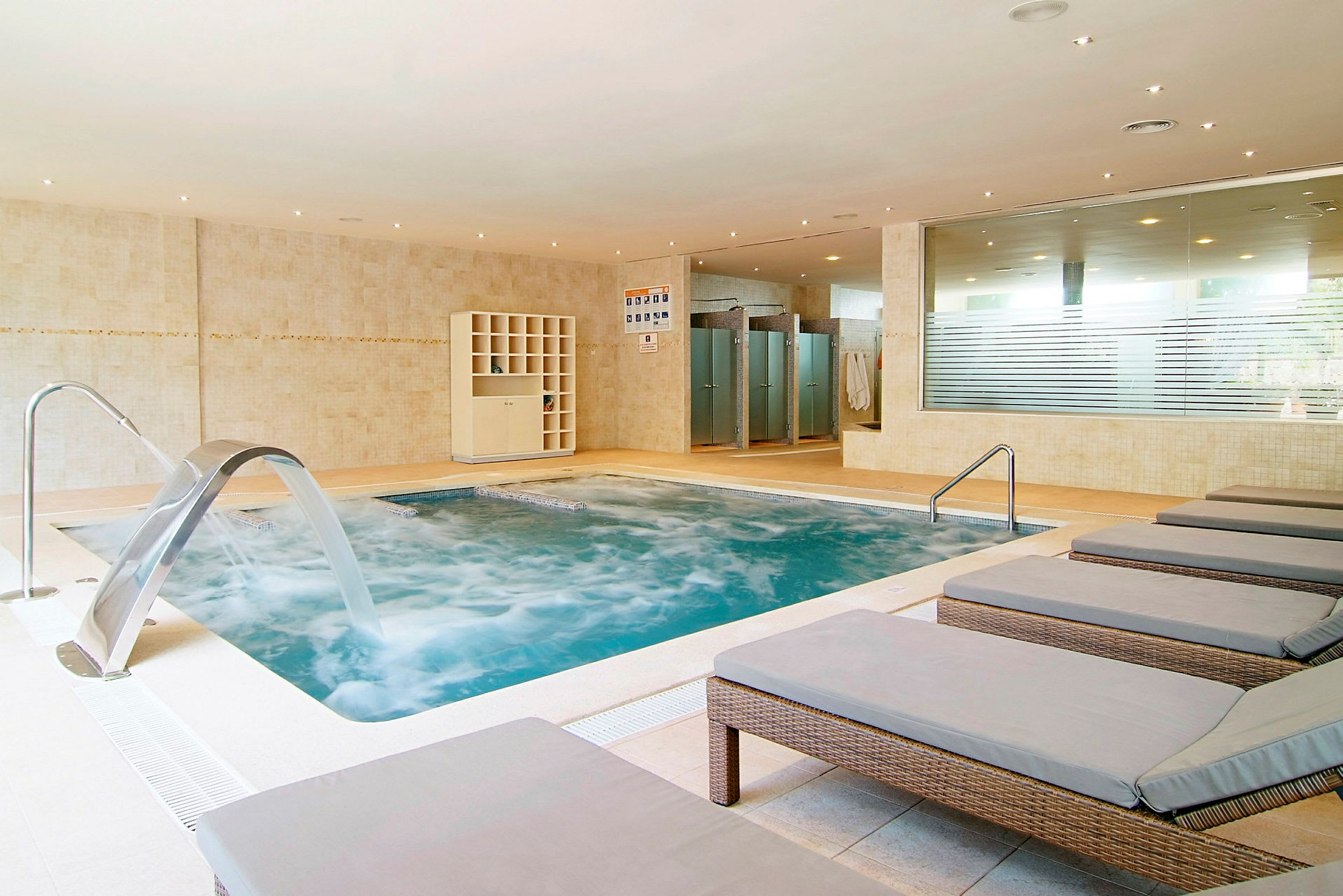En inomhuspool med jacuzzi och en fontän under ett välbelyst tak. I förgrunden finns solstolar och i bakgrunden syns duschkabiner. (AI-genererad text)