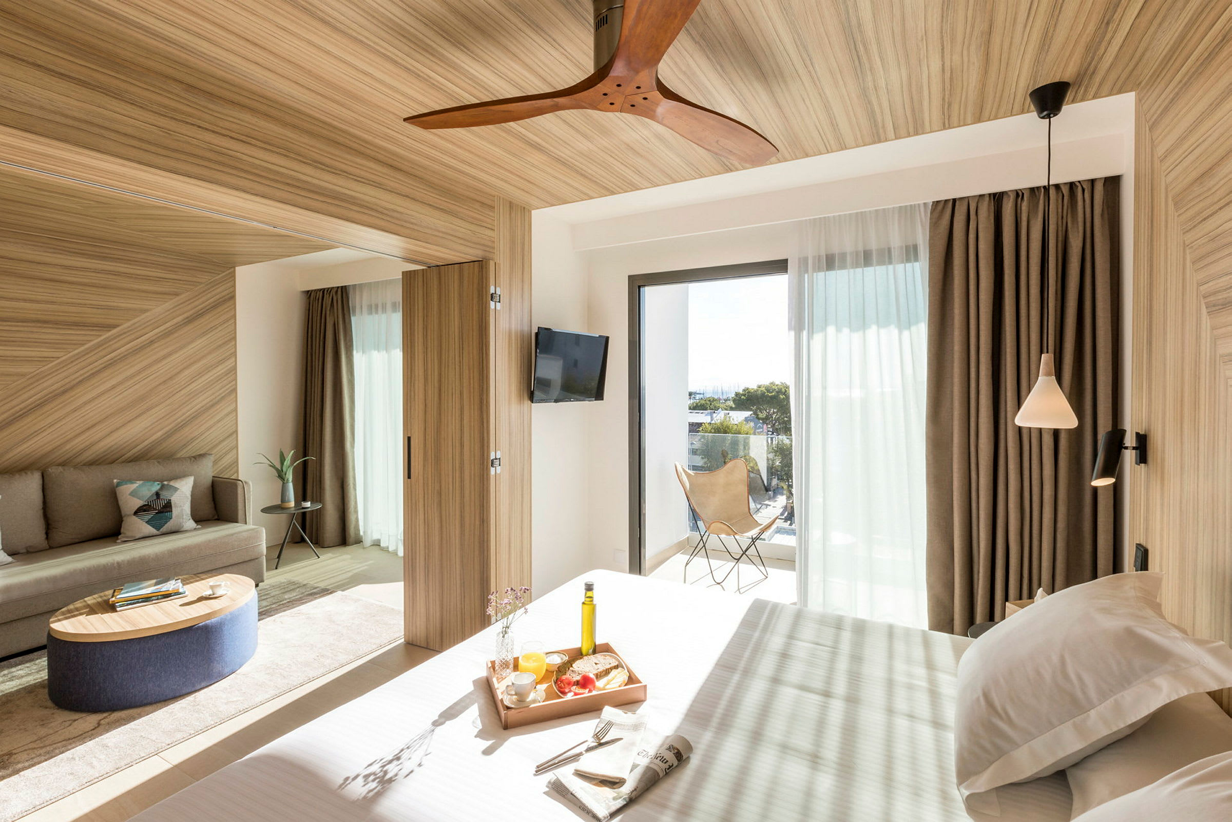 Ett modernt hotellrum med trädetaljer och stora fönster som släpper in naturligt ljus. Det finns en säng med frukostbrickor, en soffa och en balkong med utsikt. (AI-genererad text)