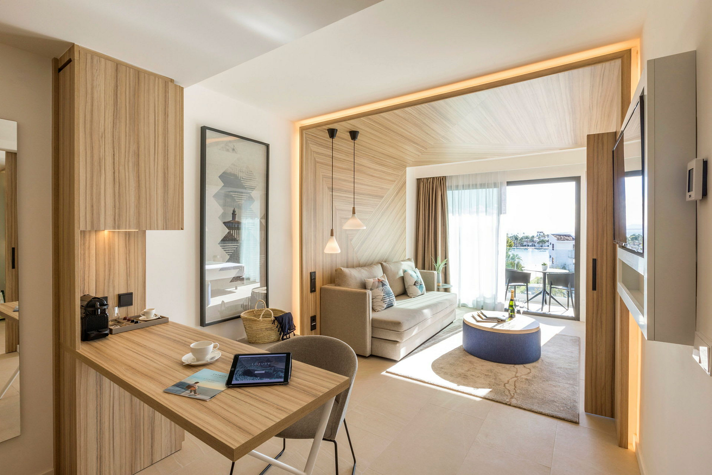 Ett modernt hotellrum med trädetaljer, en sittgrupp och ett litet bord på en matta. Balkongen har utsikt över en trädgård och vattnet utanför. (AI-genererad text)