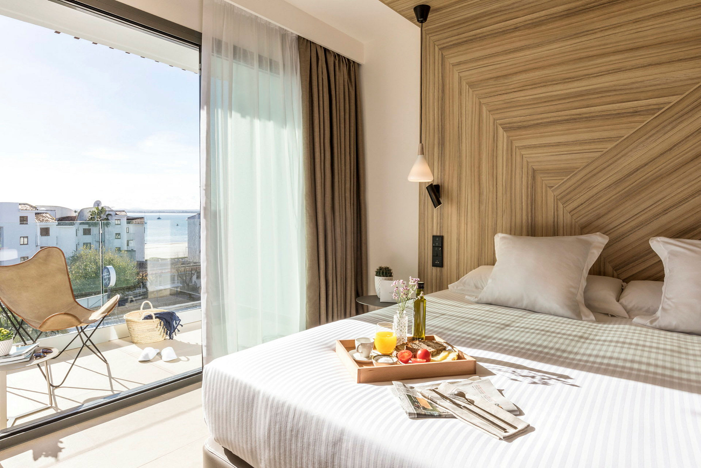 Ett ljust hotellrum med en bäddad säng med en matbricka. Stora glasdörrar leder till en balkong med en utsikt över havet. (AI-genererad text)