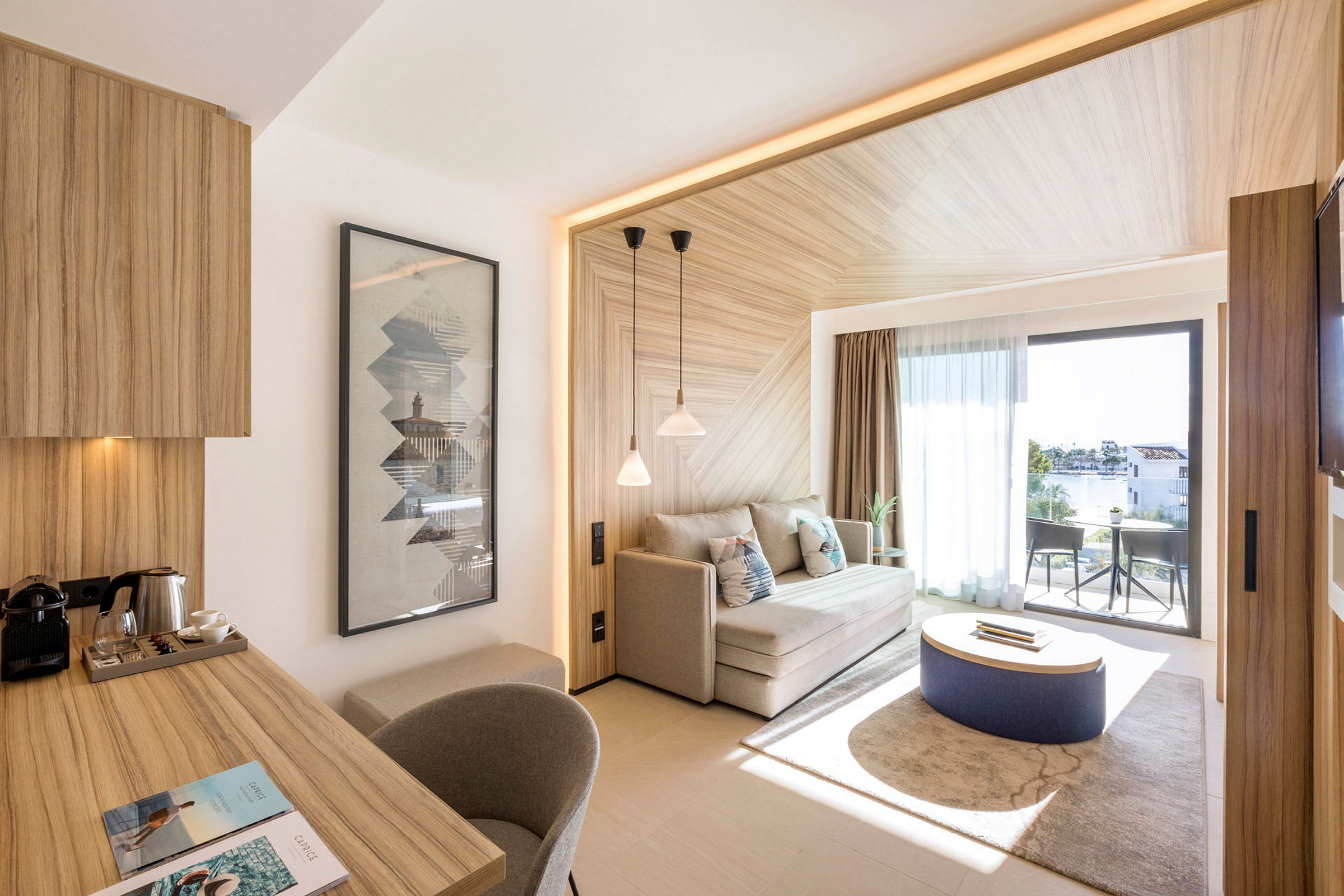 Ett modernt hotellrum med ljusa trämöbler inklusive ett skrivbord och en soffa, samt ett stort fönster som vetter mot en möblerad balkong. (AI-genererad text)