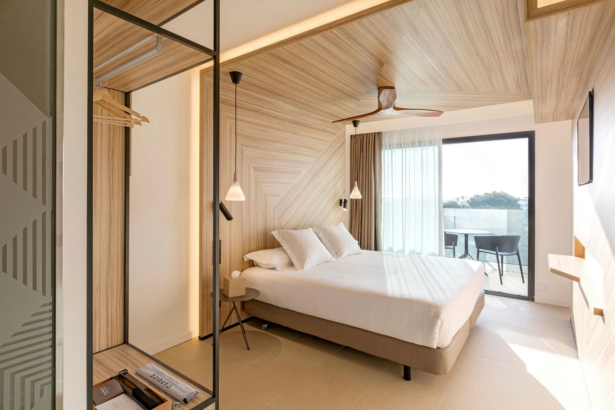 Ett hotellrum med en ljus och modern design, en säng med vita sängkläder, träinredning och ett fönster som leder till en balkong. (AI-genererad text)