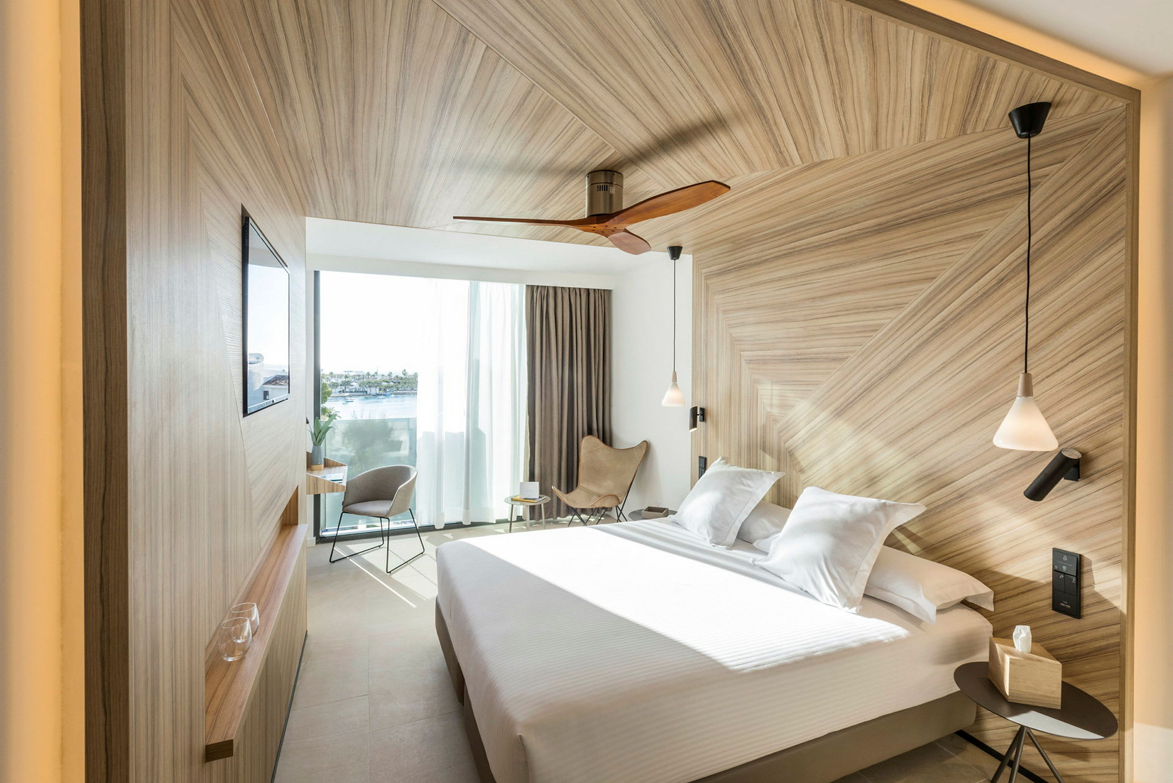 Ett modernt hotellrum med en stor säng, elegant träpanel, och stora fönster som släpper in dagsljus. (AI-genererad text)