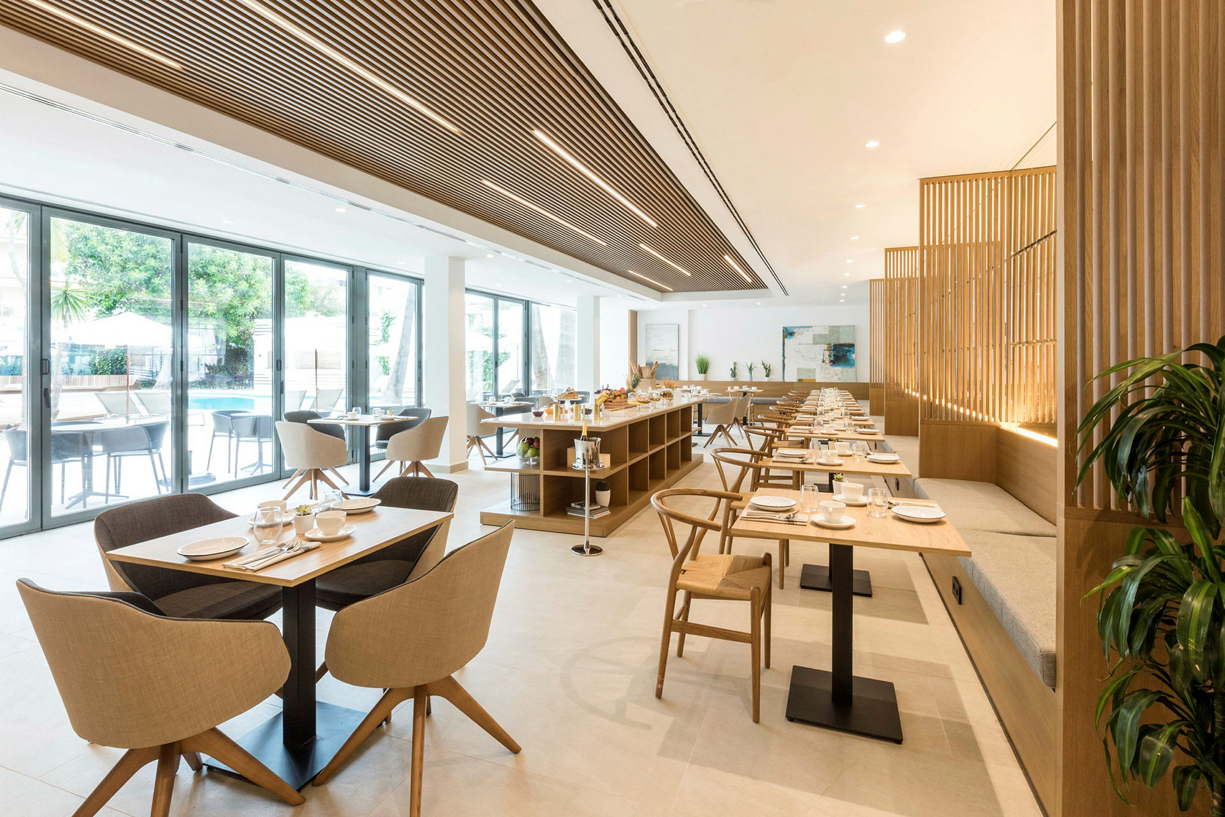 En ljus restaurang med bord och stolar i trä och textil. Glasfönster vetter mot en utomhuspool. (AI-genererad text)