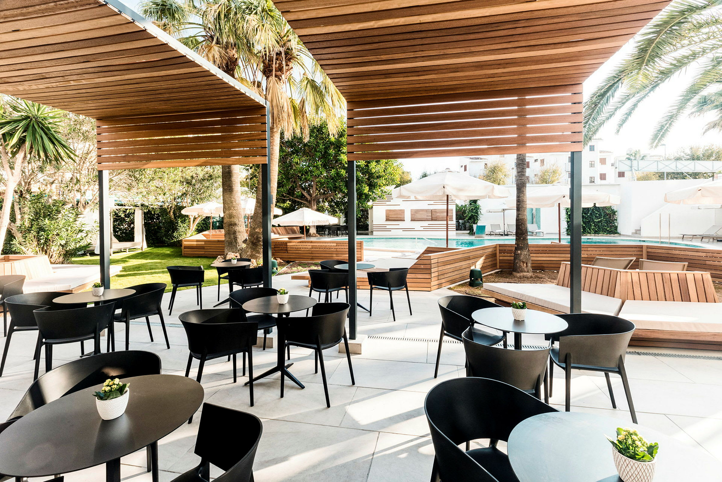 Utomhusområde med sittgrupper av bord och stolar under träpergola, omgiven av grönska och palmer. I bakgrunden syns en pool och solstolar med parasoller. (AI-genererad text)
