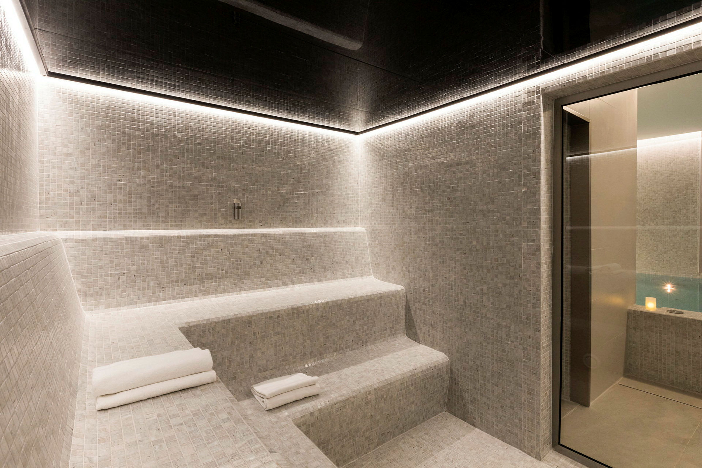 Bild på ett modernt hamam med mosaikklädda sittplatser och en glaskant som leder till en pool. (AI-genererad text)