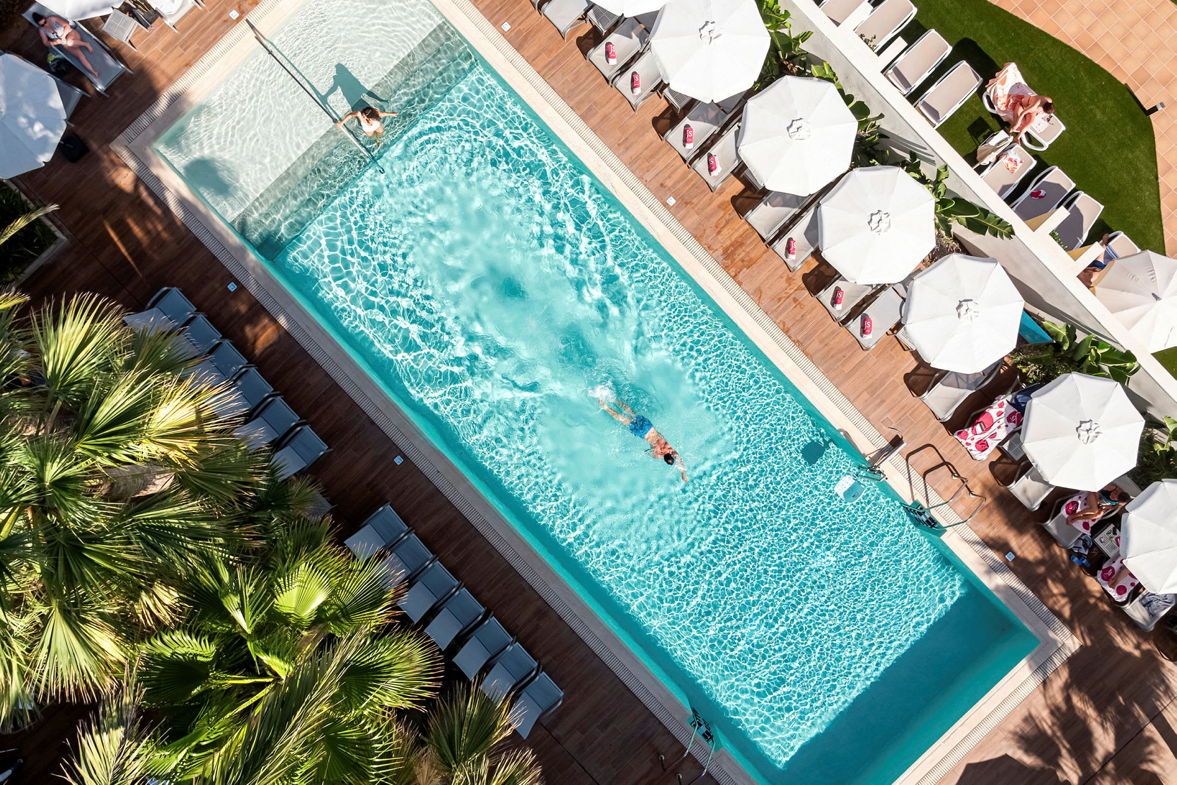 En fågelperspektivbild av en swimmingpool med en simmande person och solstolar med parasoller runtom. (AI-genererad text)