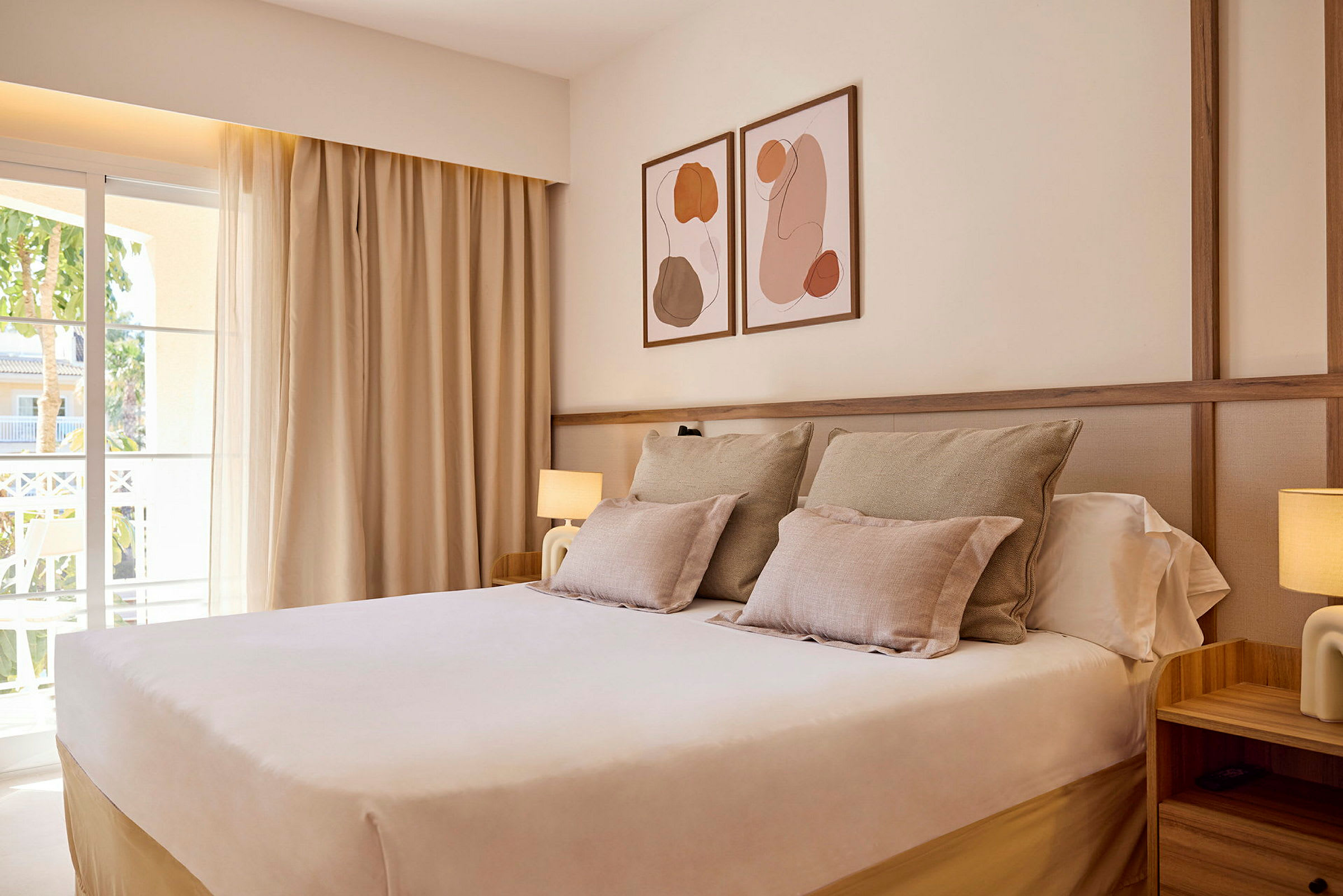 Ett hotellrum med en stor säng, dekorerad med ljusa sängkläder och kuddar. Tavlor hänger på väggen ovanför sängen. Ett fönster med draperier syns till vänster. (AI-genererad text)
