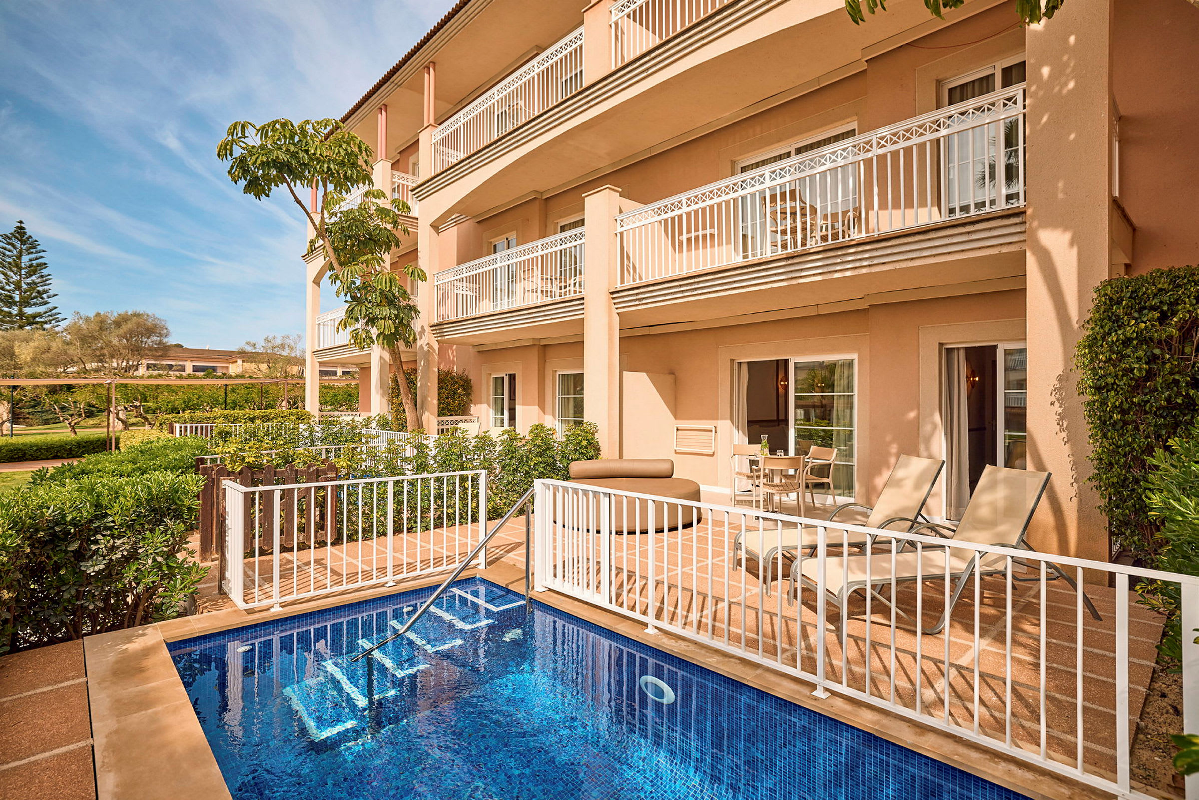 Byggnad med balkonger, pool och terrass med solstolar och växter i förgrunden. (AI-genererad text)