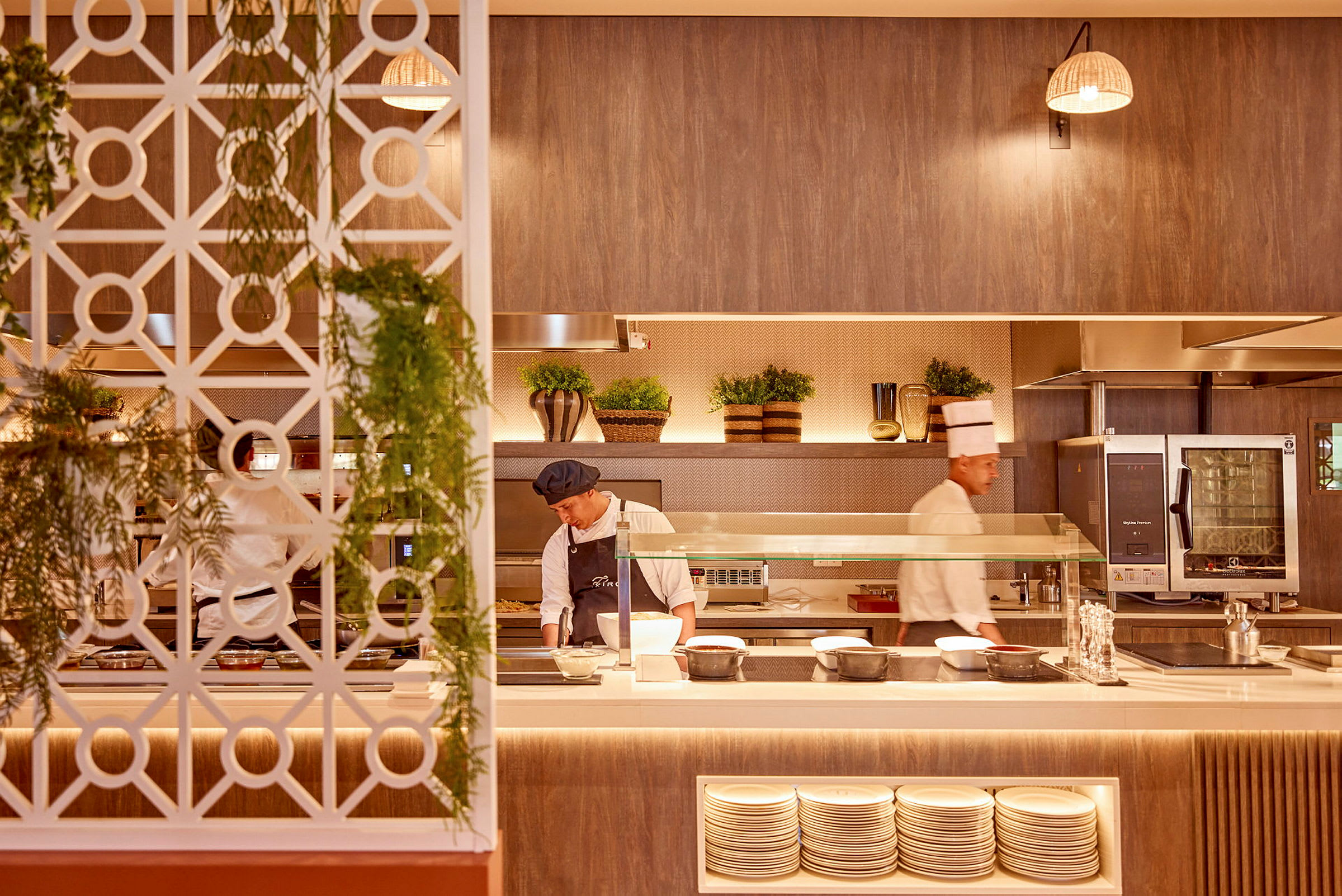 En restaurangkök med kockar som arbetar, en dekorativ vägg med hängande grönska, tallrikar i hyllor och inredning med rustika detaljer. (AI-genererad text)