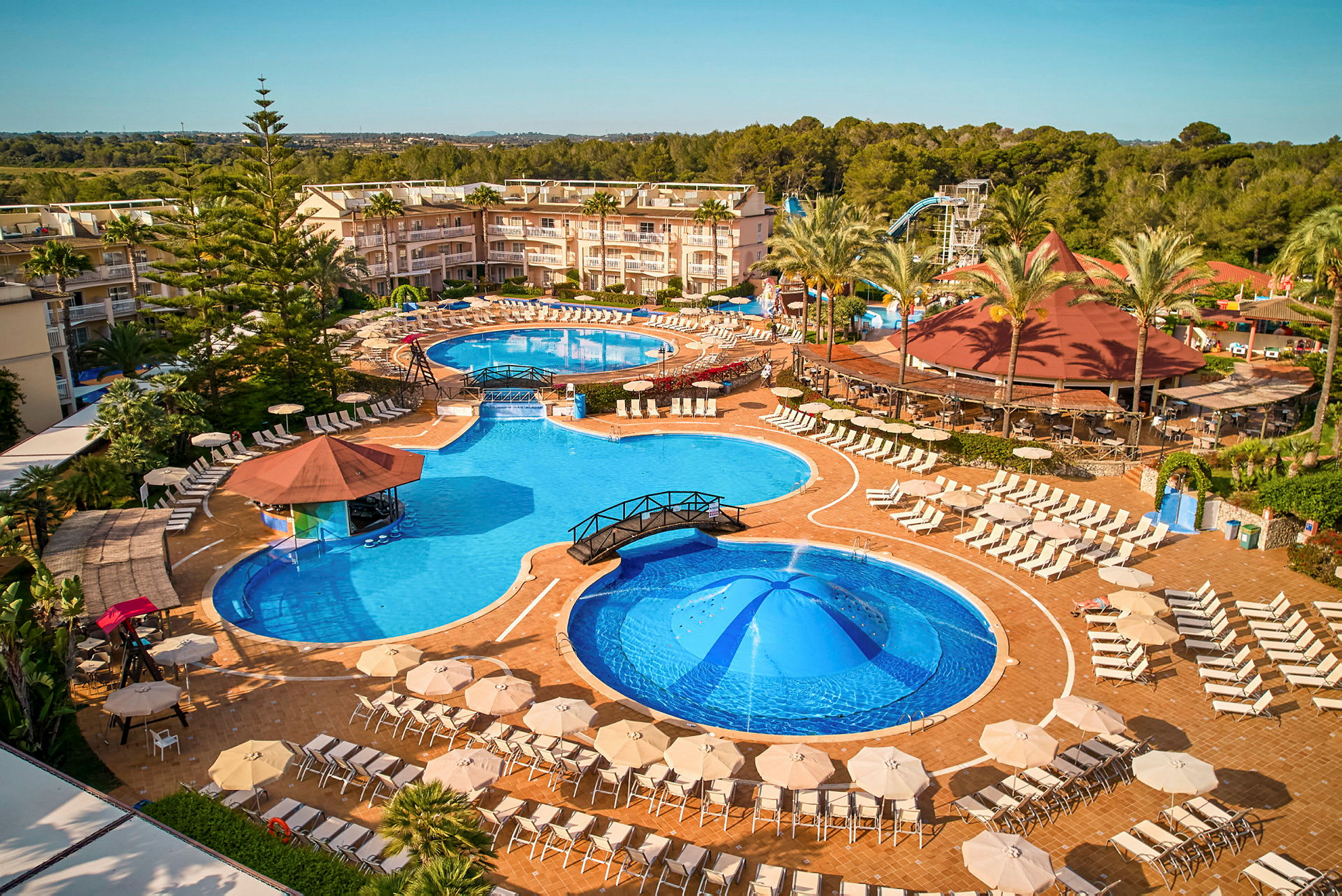 En stor resort med flera pooler, parasoller och solstolar omgiven av palmer och grönska. (AI-genererad text)