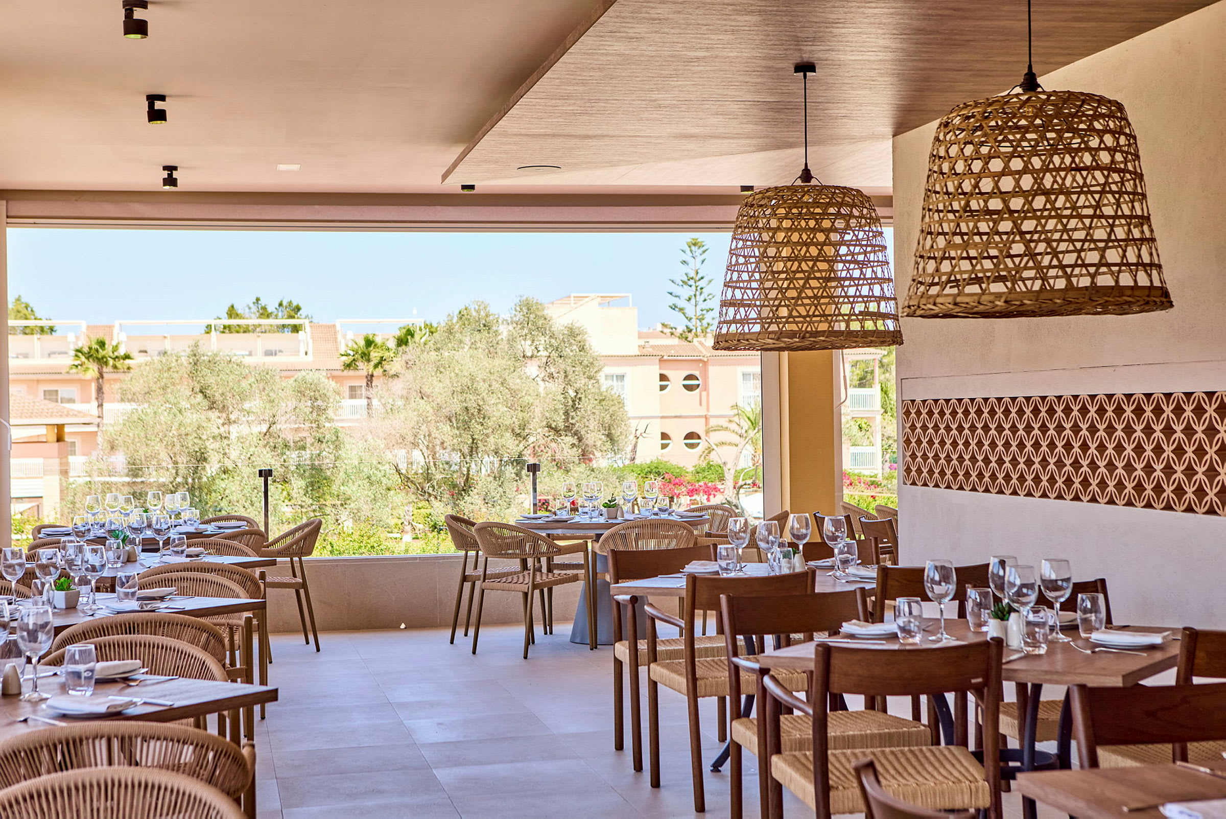 En restaurang med dukade bord och stolar, utsmyckad med flätade ljuslampor, och utsikt över en trädgård och byggnader. (AI-genererad text)