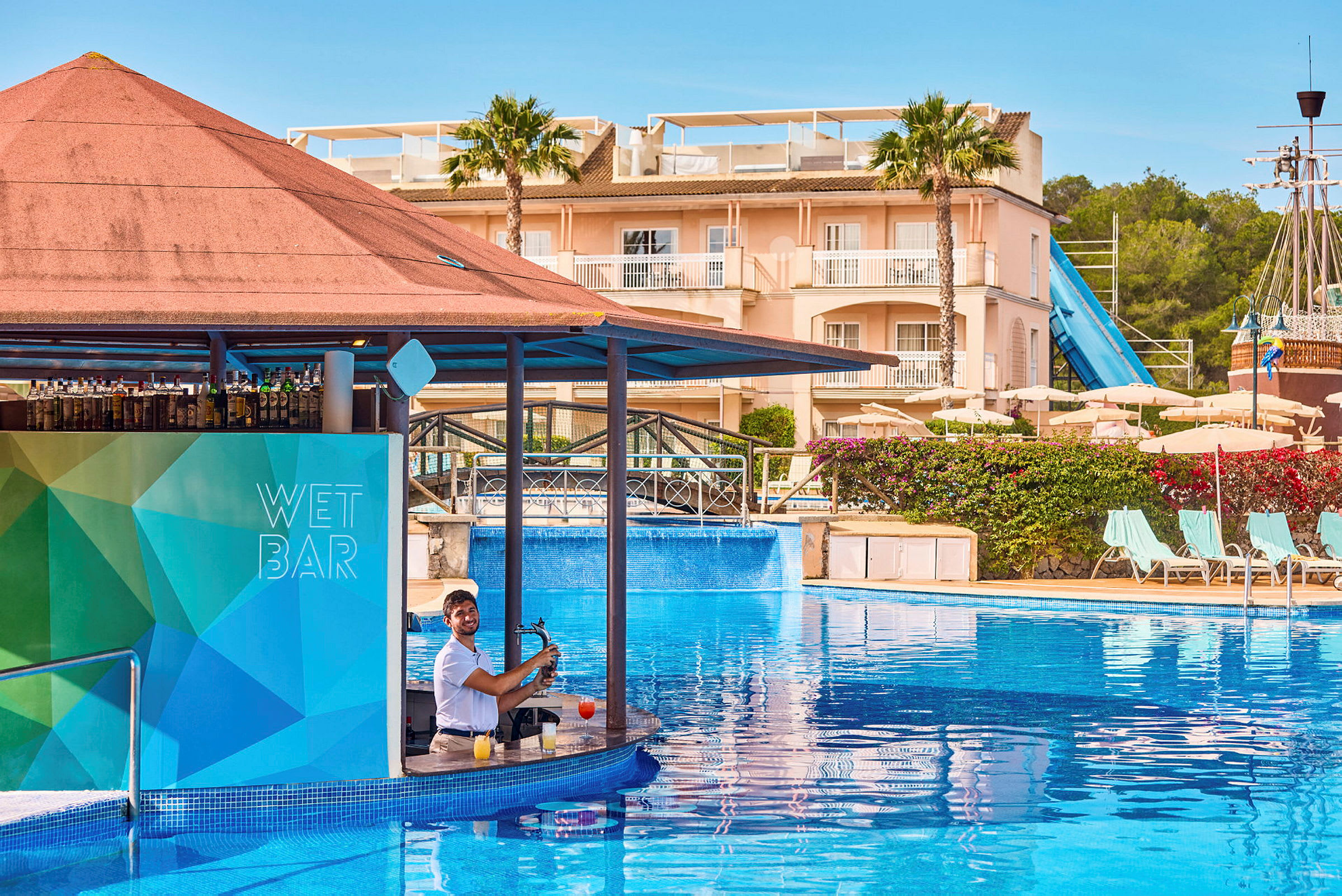 Bar i en swimmingpool med en bartender. I bakgrunden syns en byggnad, solstolar, parasoller och en vattenrutschkana. (AI-genererad text)