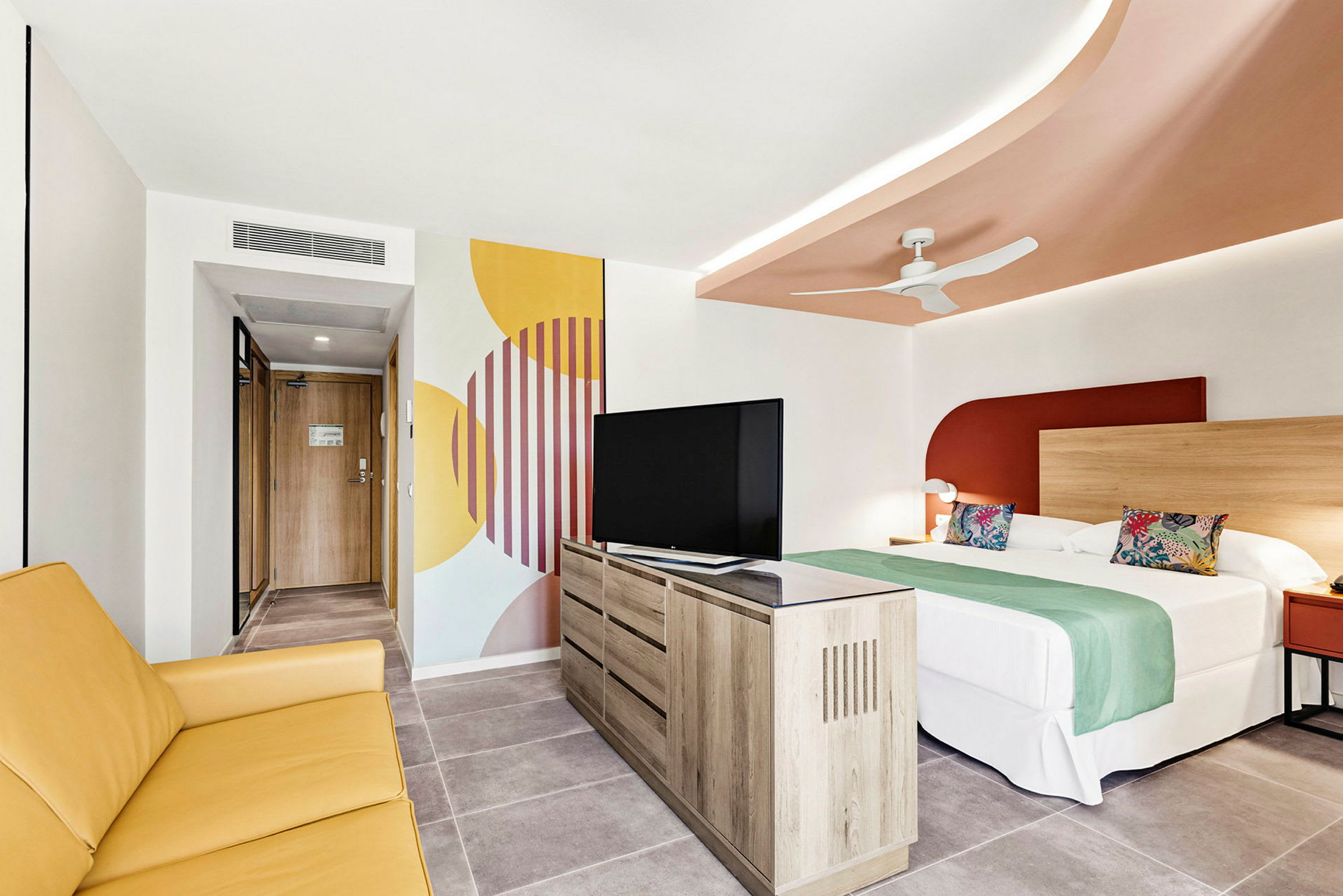 Hotellrum med en säng, gul soffa, TV och dekorativ väggmålning. (AI-genererad text)