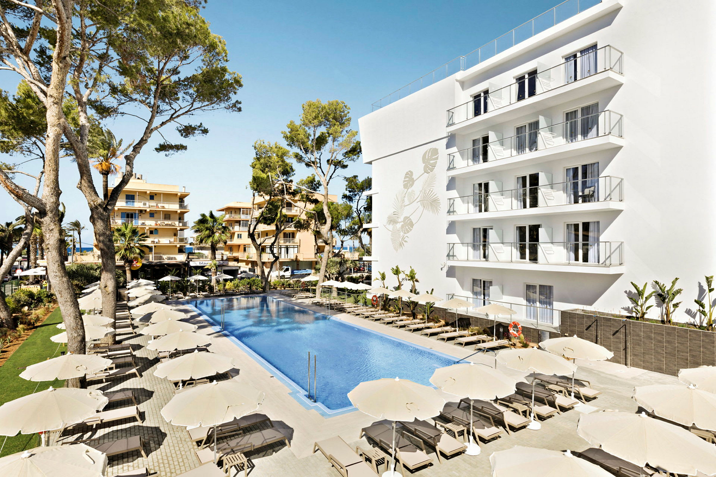 Bild av ett hotellområde med en stor pool omgiven av solstolar och parasoller. En vit byggnad med flera balkonger samt träd i bakgrunden. (AI-genererad text)