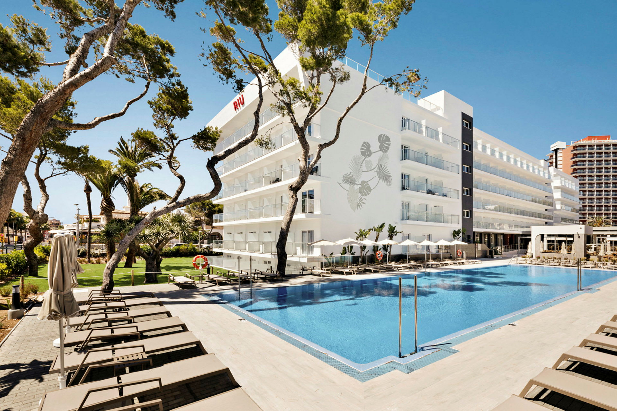 Ett hotell med en pool i förgrunden och solstolar runt omkring, omgiven av träd och blå himmel. (AI-genererad text)