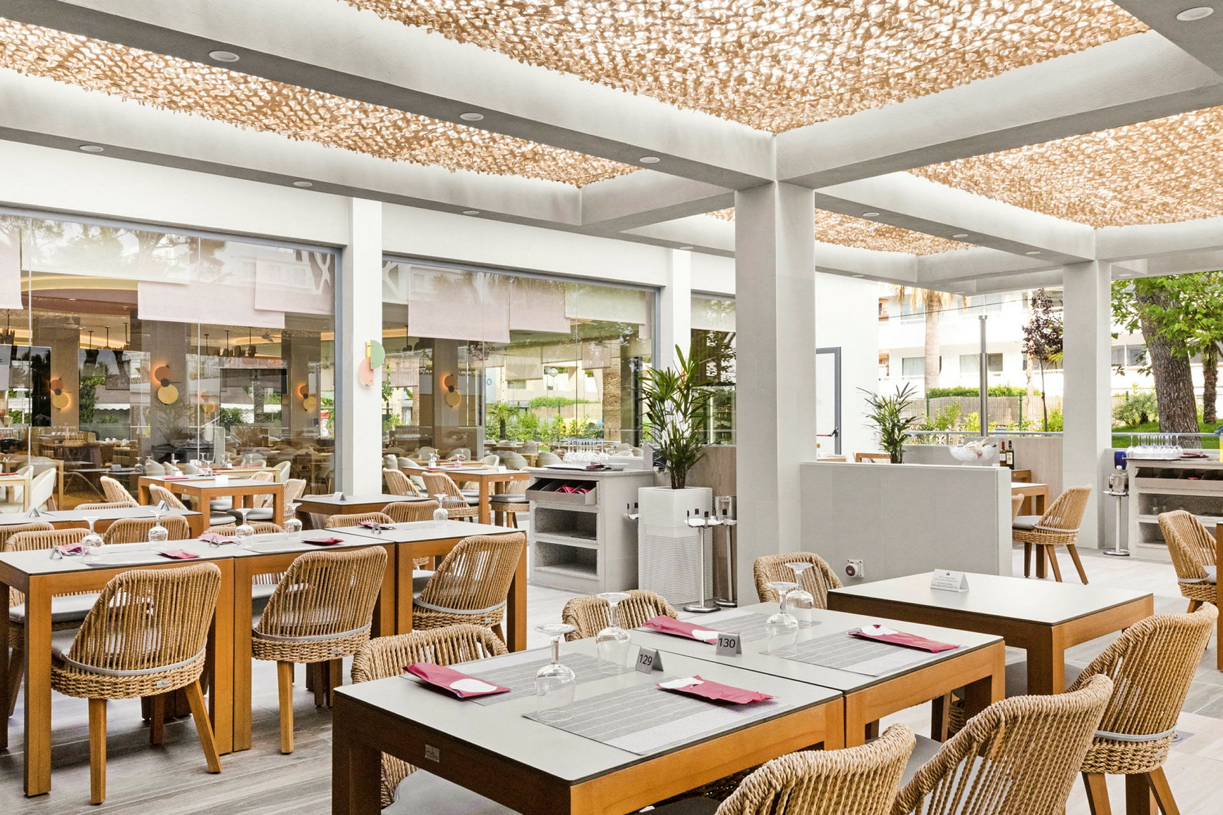 En ljus restaurang med träbord och rottingstolar, placerad i en modern inomhusmiljö med ett dekorativt tak och glasväggar som öppnar upp mot utsidan. (AI-genererad text)
