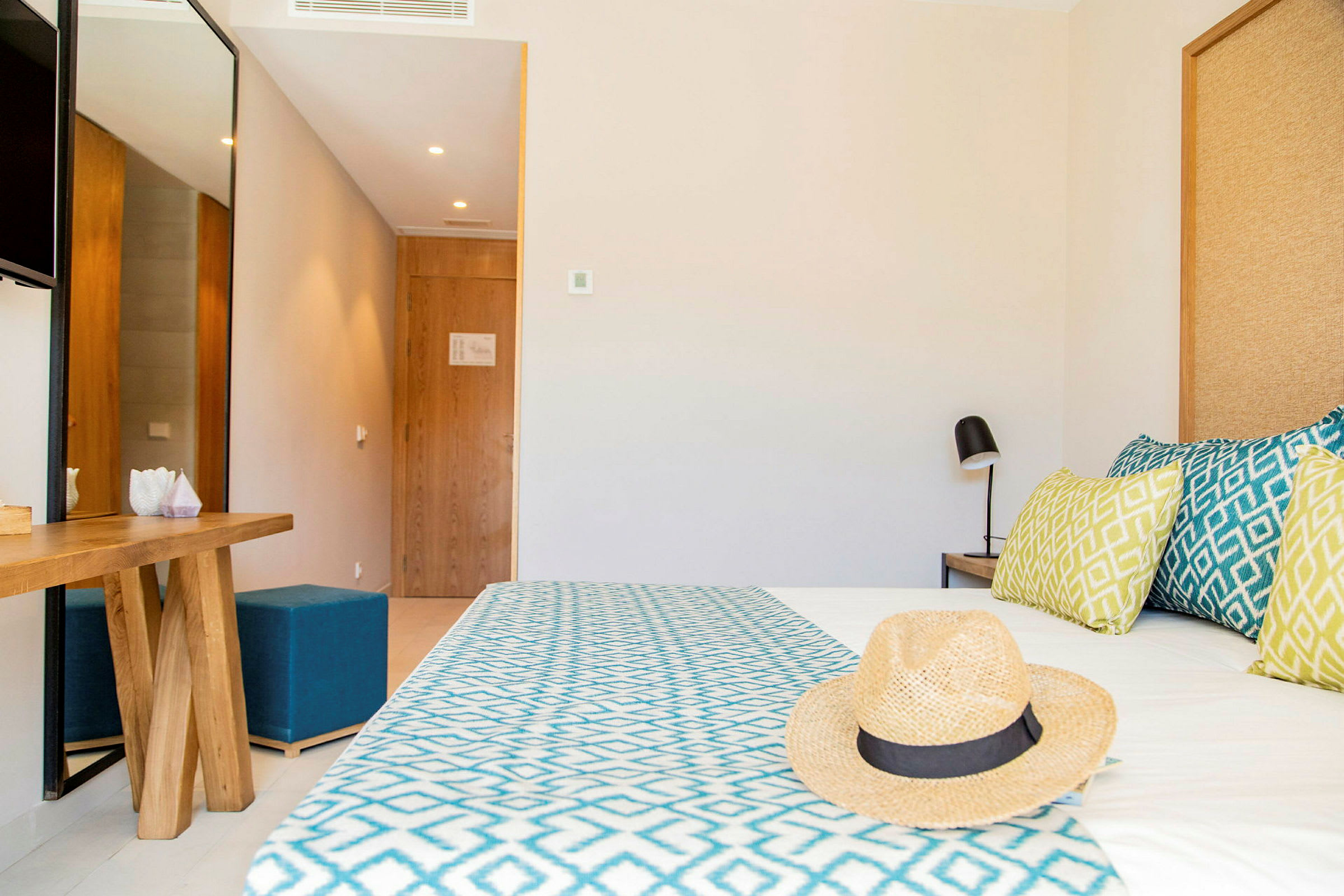 Ett hotellrum med en säng med mönstrade kuddar, en halmhatt ligger på sängen, och en spegel och träbord är synliga. (AI-genererad text)