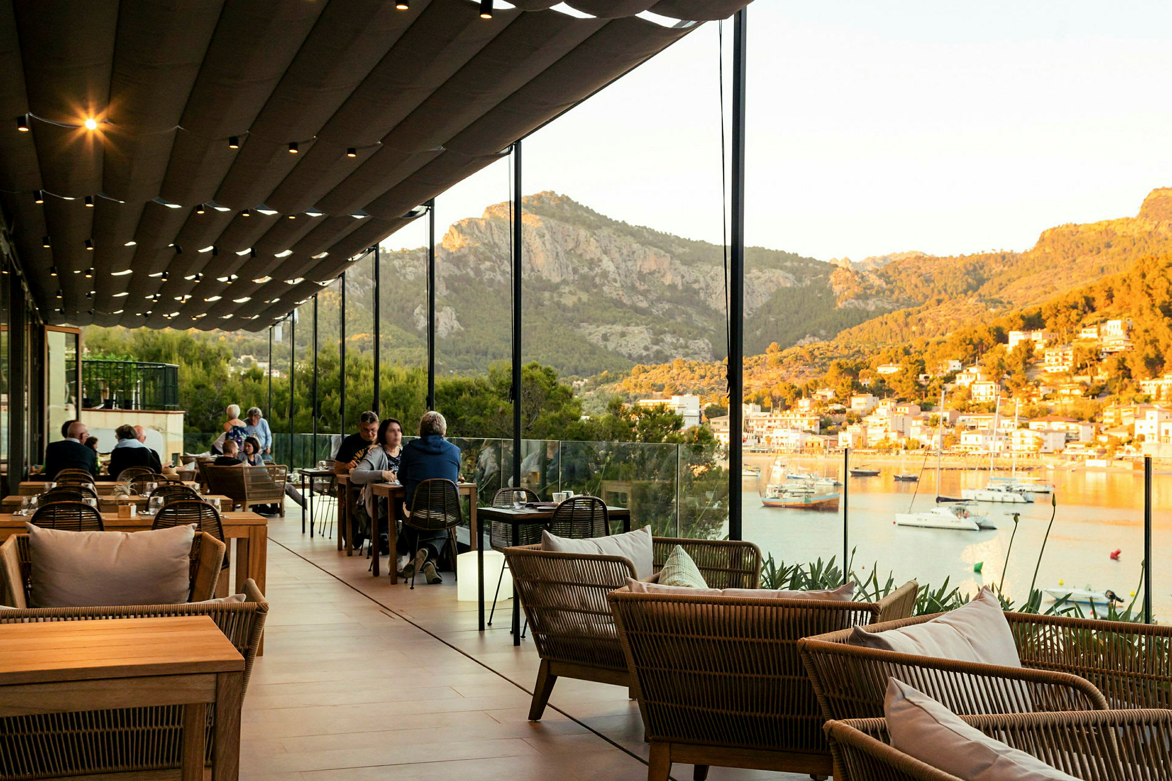 En restaurang med sittplatser utomhus under ett täcke med utsikt över en hamn och berg i bakgrunden. (AI-genererad text)
